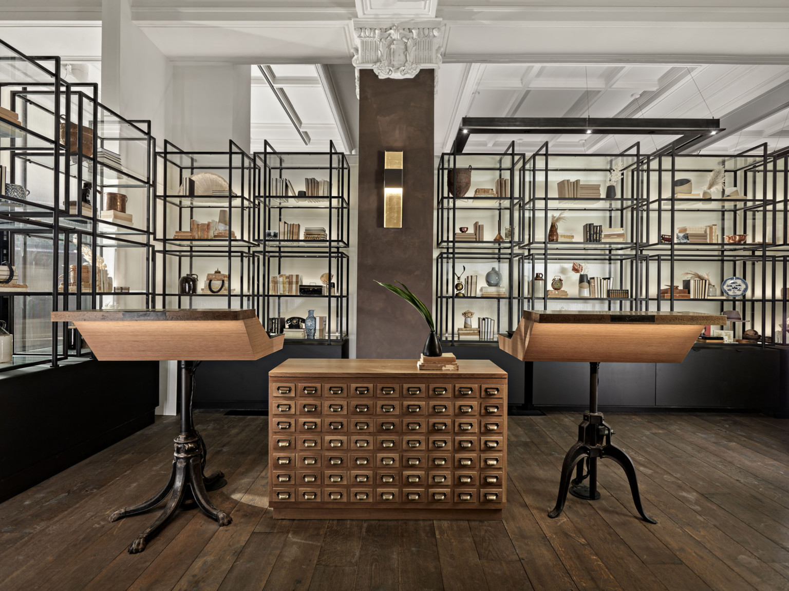 Sloping wood tables with dark metal legs separated by table with small drawers. Behind, glass and metal shelves of items