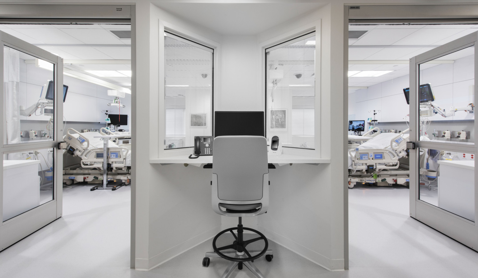 Desk with view into 2 hospital rooms, each with beds and screens. The white desk fits into a triangular space between doors