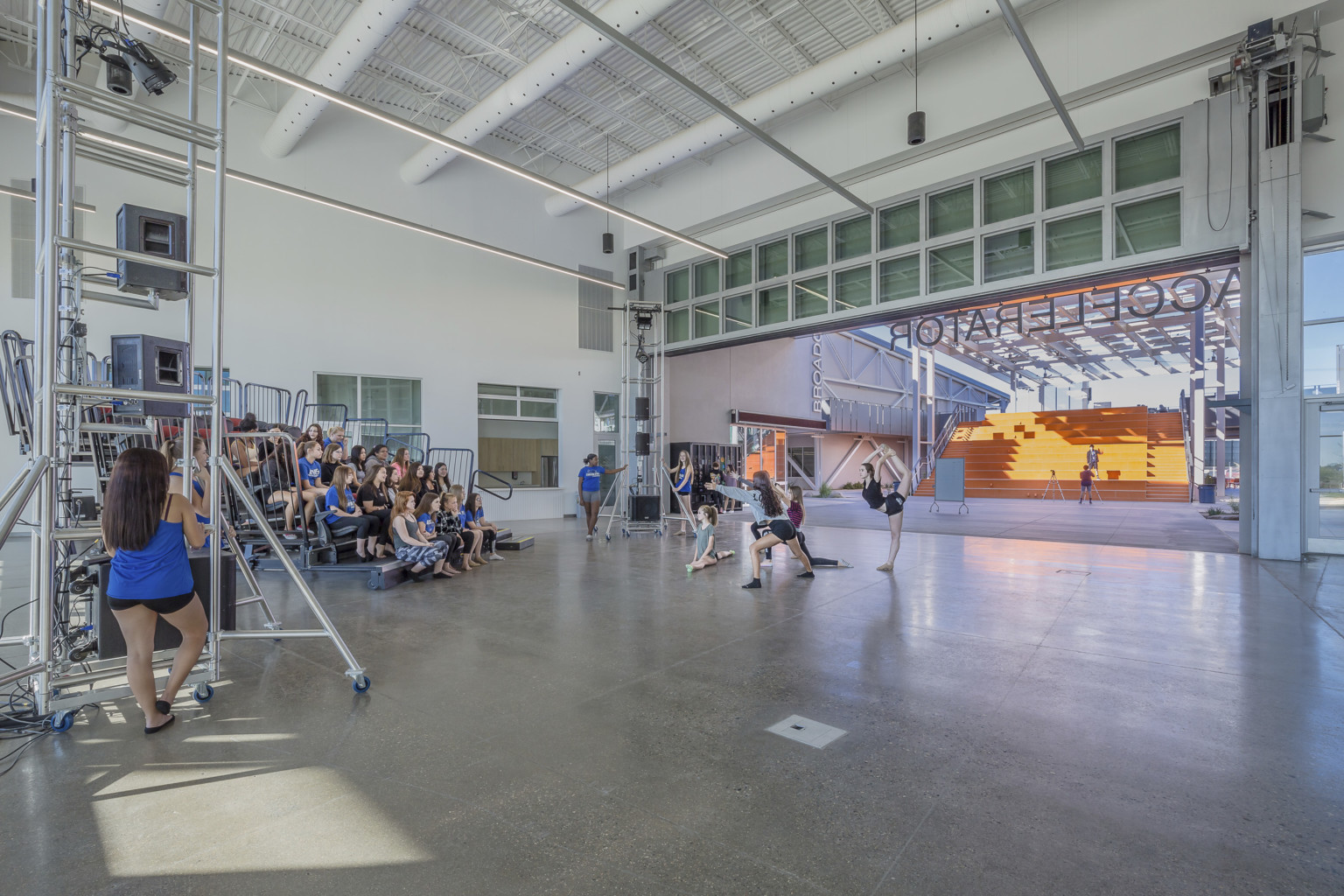 A white room, with the wall opened facing orange stairs, has portable bleachers and equipment