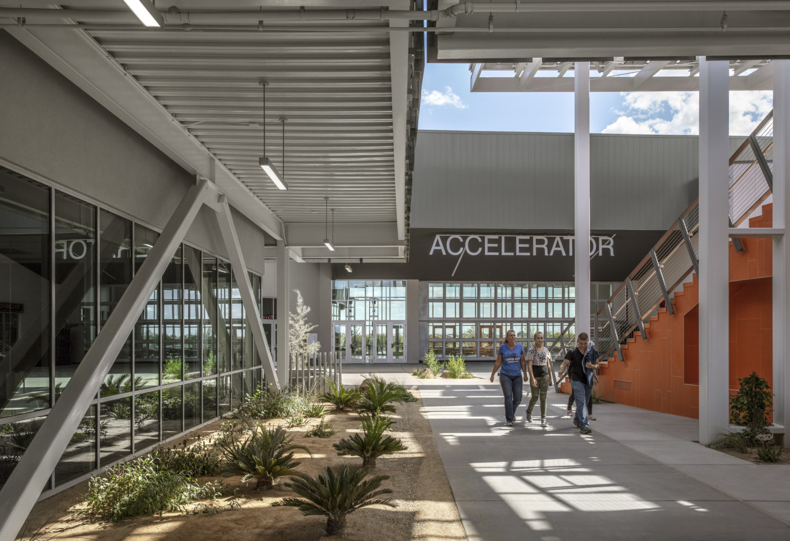 Breezeway to the left of the orange stairs. Behind is a building with a dark roof and parallel walls of double height windows