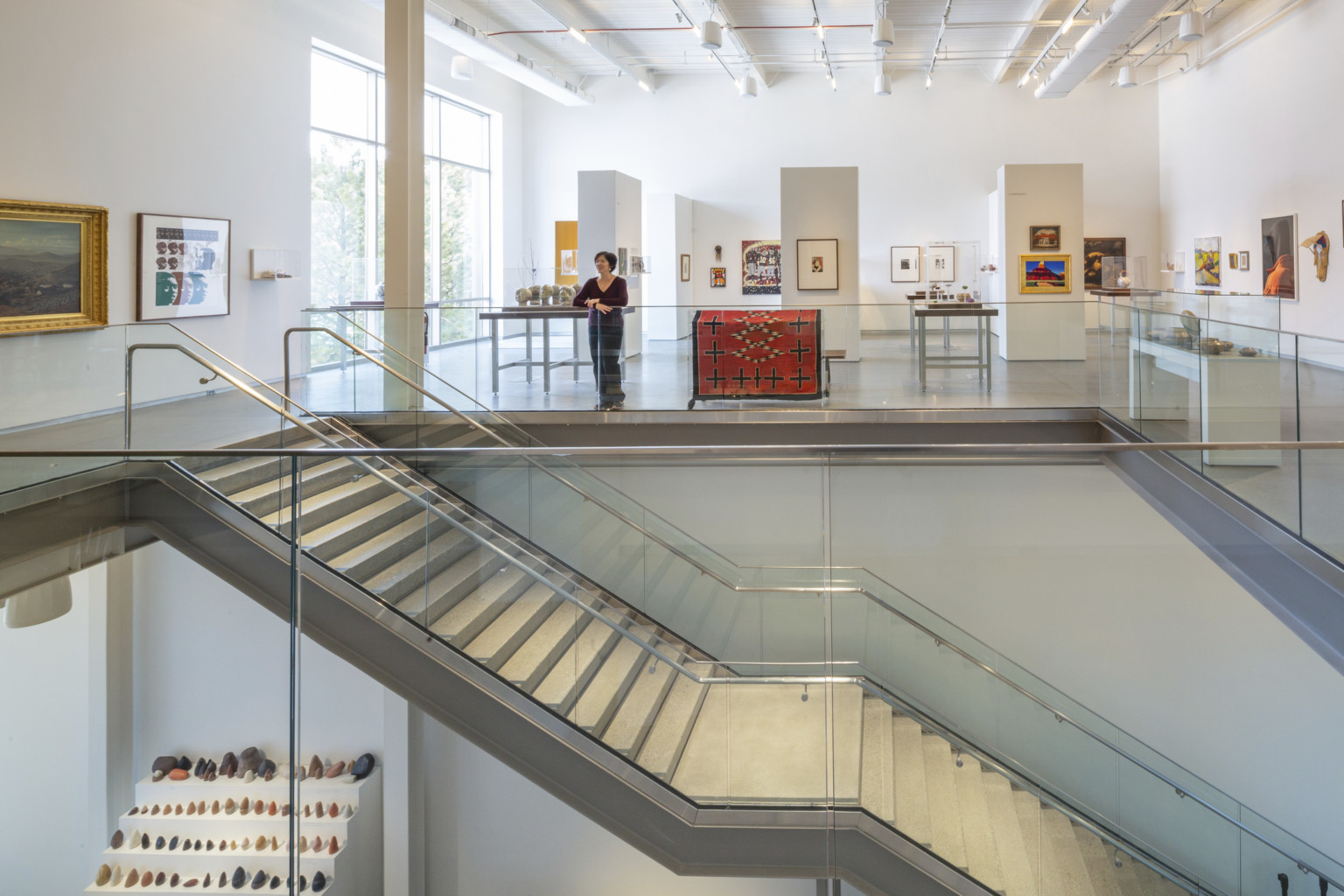 view from second story of art gallery split by staircase with white walls and custom lighting solution illuminating exhibits