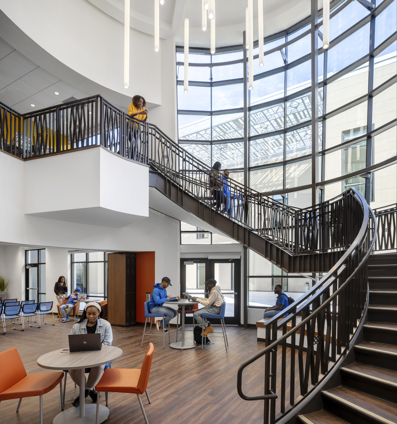 Curved staircase along double height windows. Above, cylindrical white pendant lights, seating area at the base of the stairs