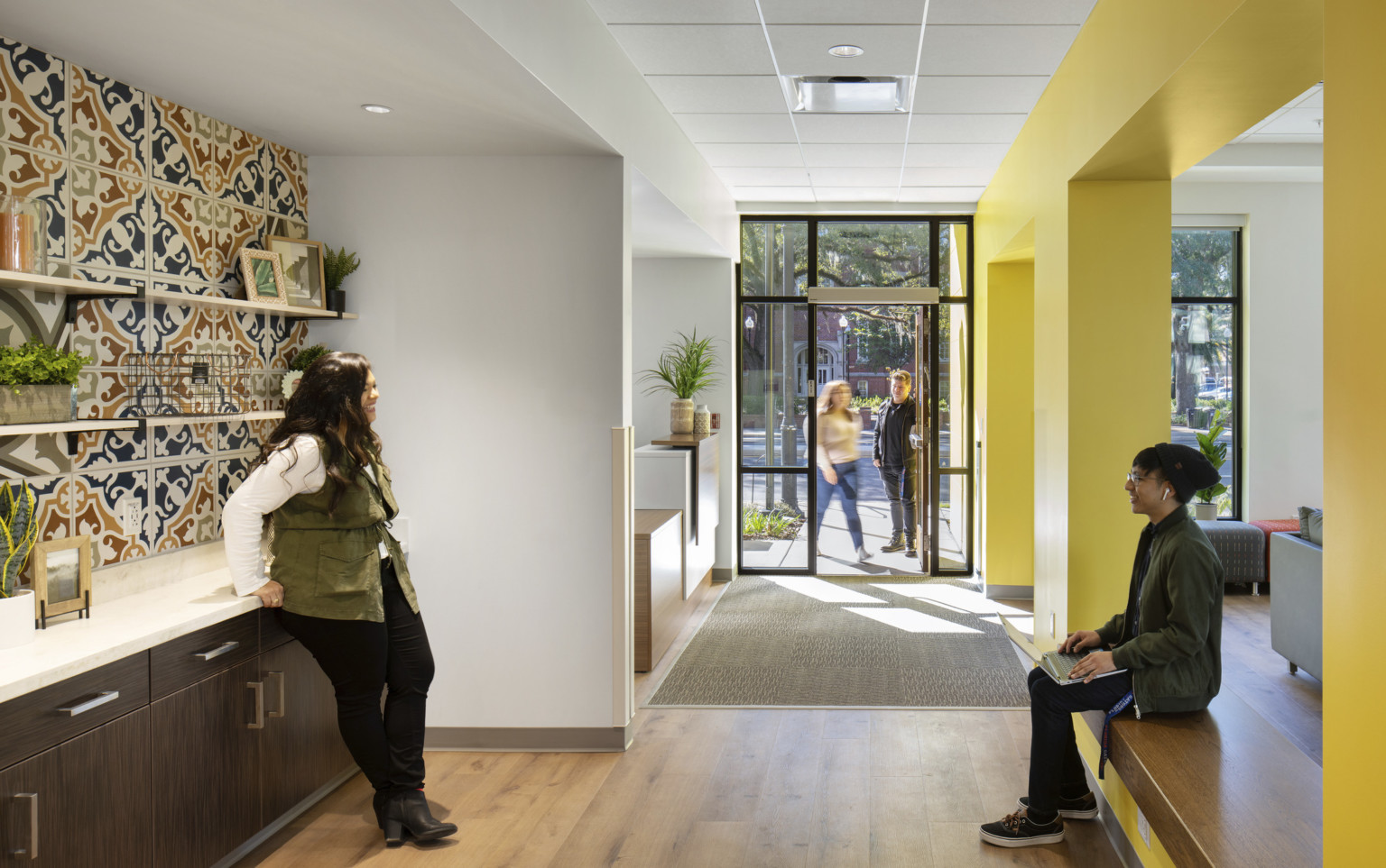 Hallway left of yellow wall to entry. White room towards the door behind recessed dark wood counter with muraled backsplash