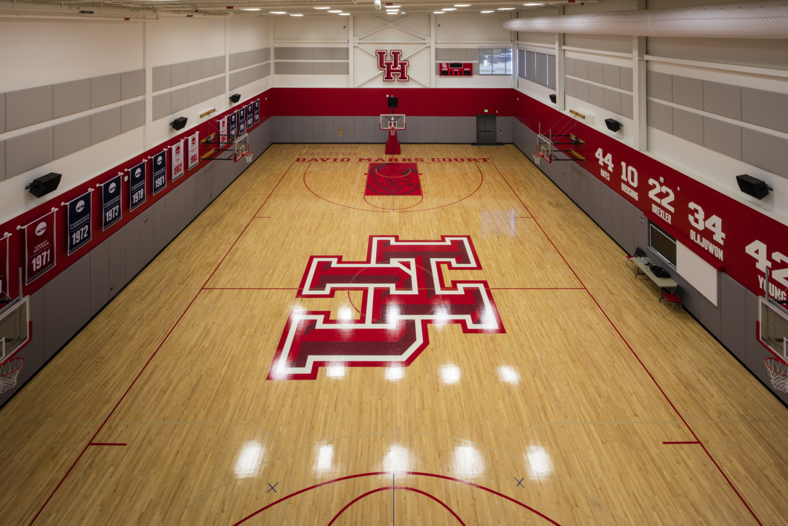 Wood practice court with basketball hoops along sides and UH logo at center. Grey and red stripes on walls with banners left