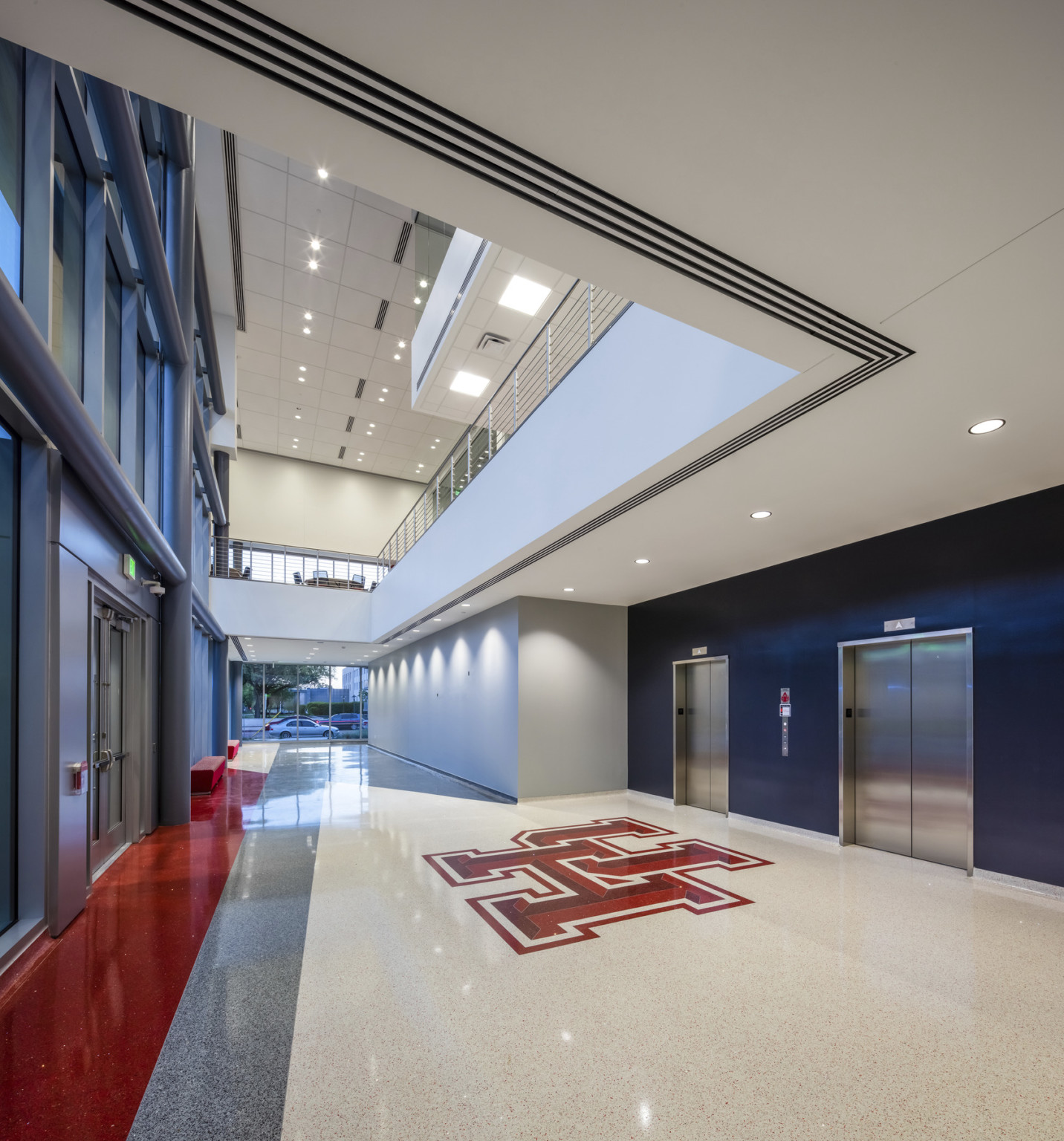 Triple height hallway with blue elevator shaft, right, recessed under white 2nd floor walkway. Large windows on far wall