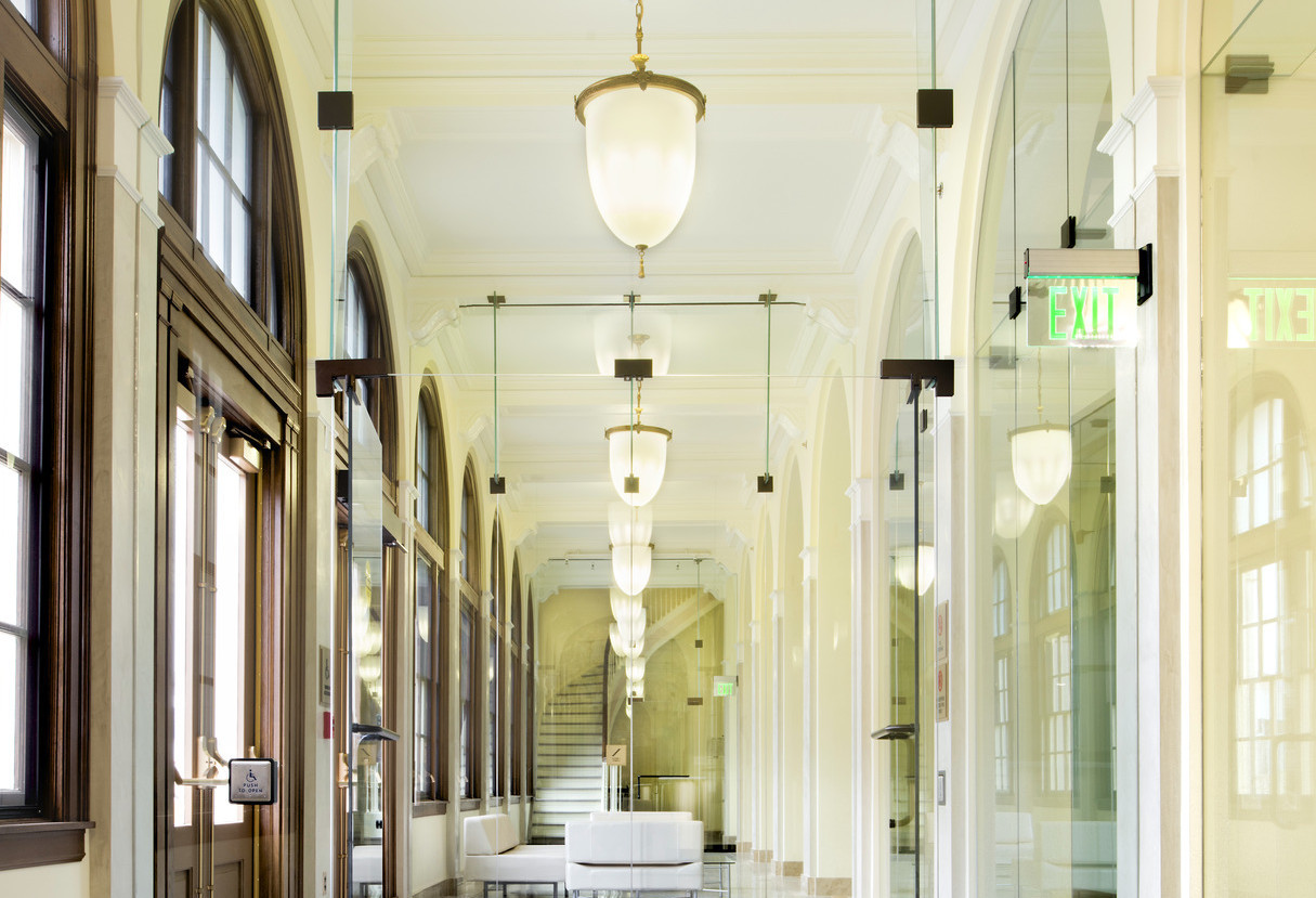 A hallway lined with arched windows and mirrors and central pendant lights leading to a curved stairwell