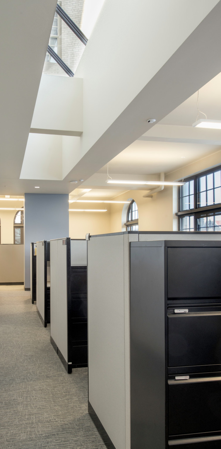 Light coming in large rectangular and arch shaped windows over a line a cubicles in a cream colored office with white ceiling