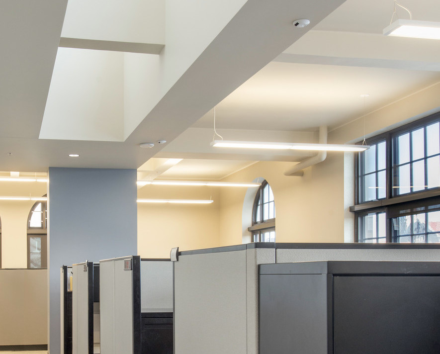Light coming in large rectangular and arch shaped windows over a line a cubicles in a cream colored office with white ceiling