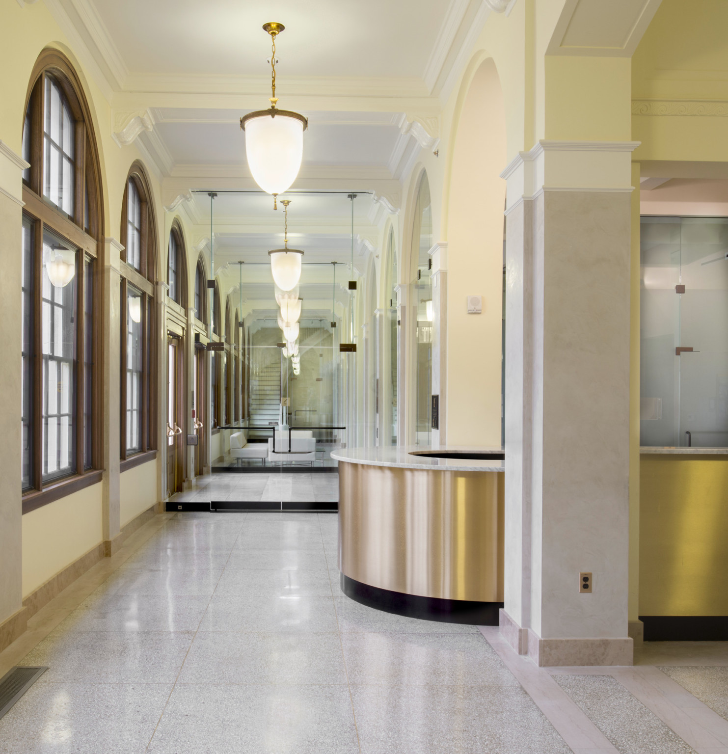 Gold reception desk with marble counter in front of a mirrored hallway with arched windows and hanging light