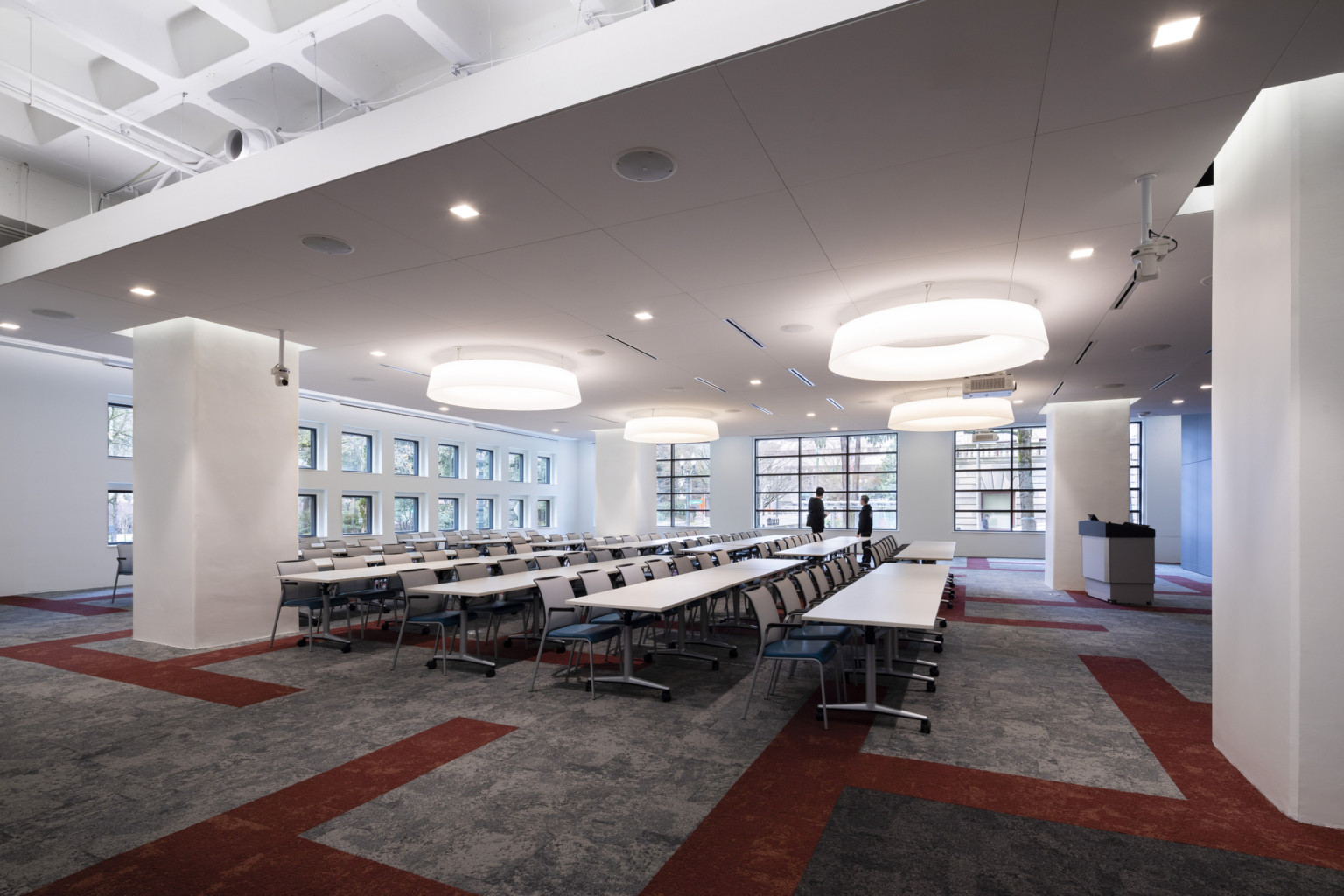 Large event space with a series of square windows and circular light fixtures contains movable tables and chairs facing podium