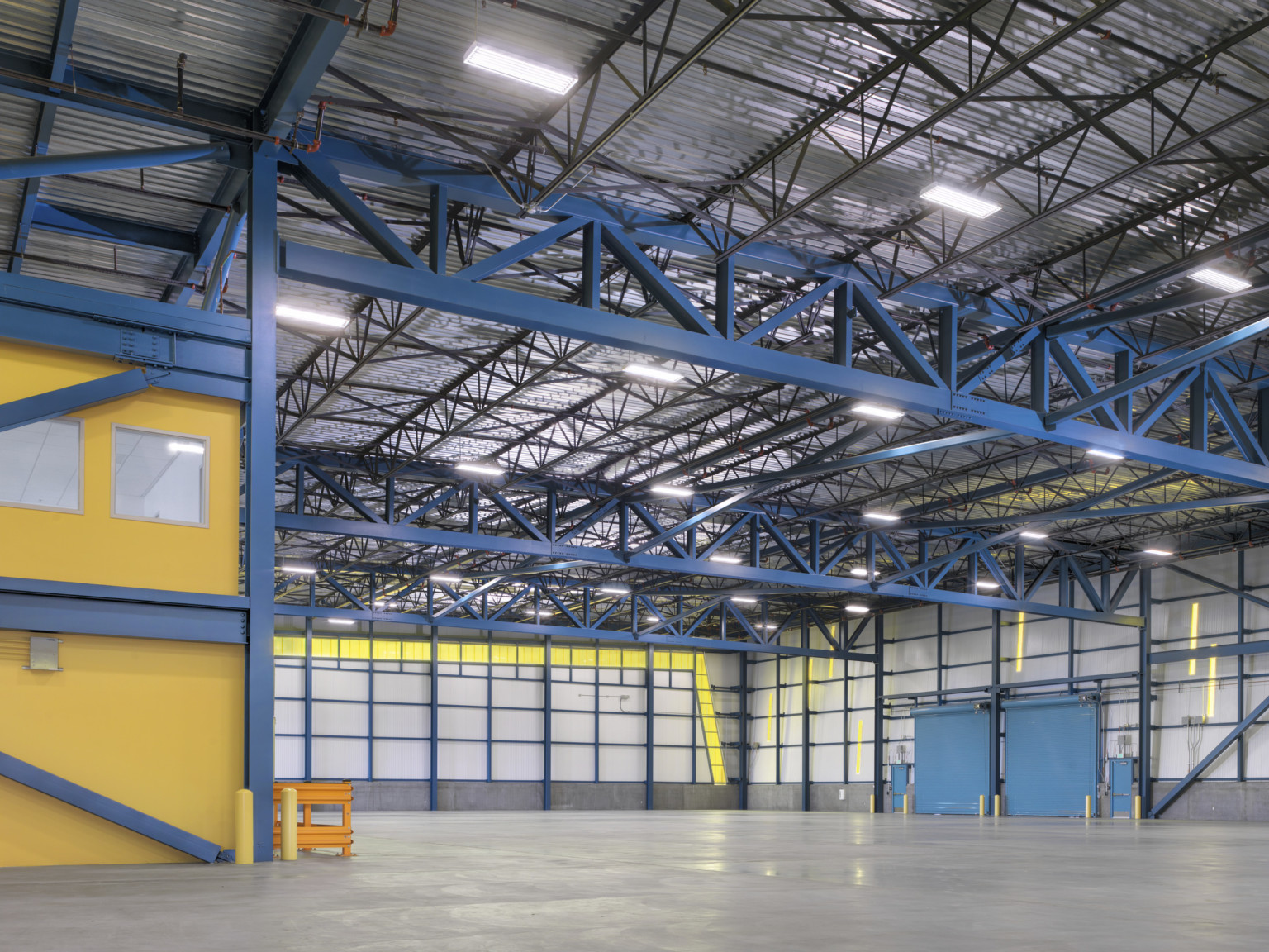 A large grey open interior with blue exposed beams and a yellow structure, left. Garage style doors on right wall, closed