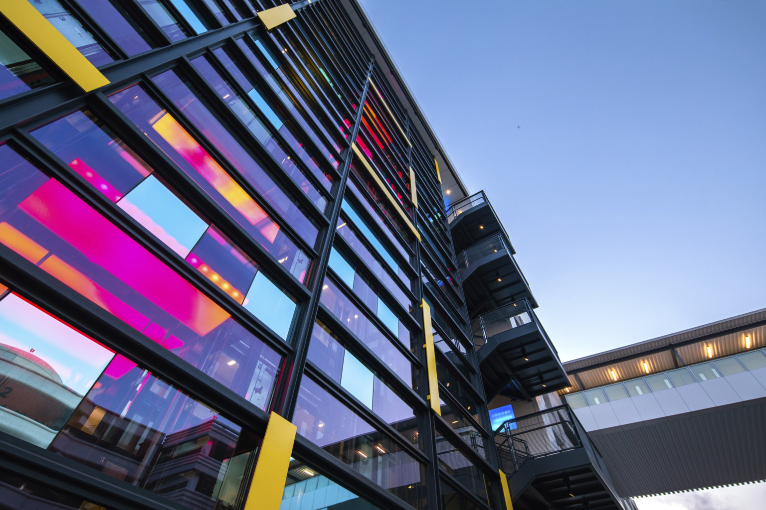 View looking up at central glass tower with iridescent window panels and staircase to the right. Skywalk over road to right