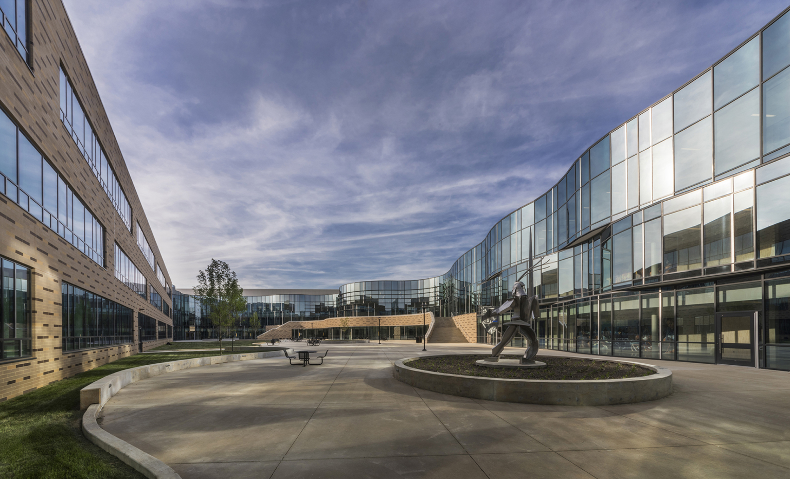 a brick building and mirrored glass facade surround a concrete courtyard with a metal sculpture