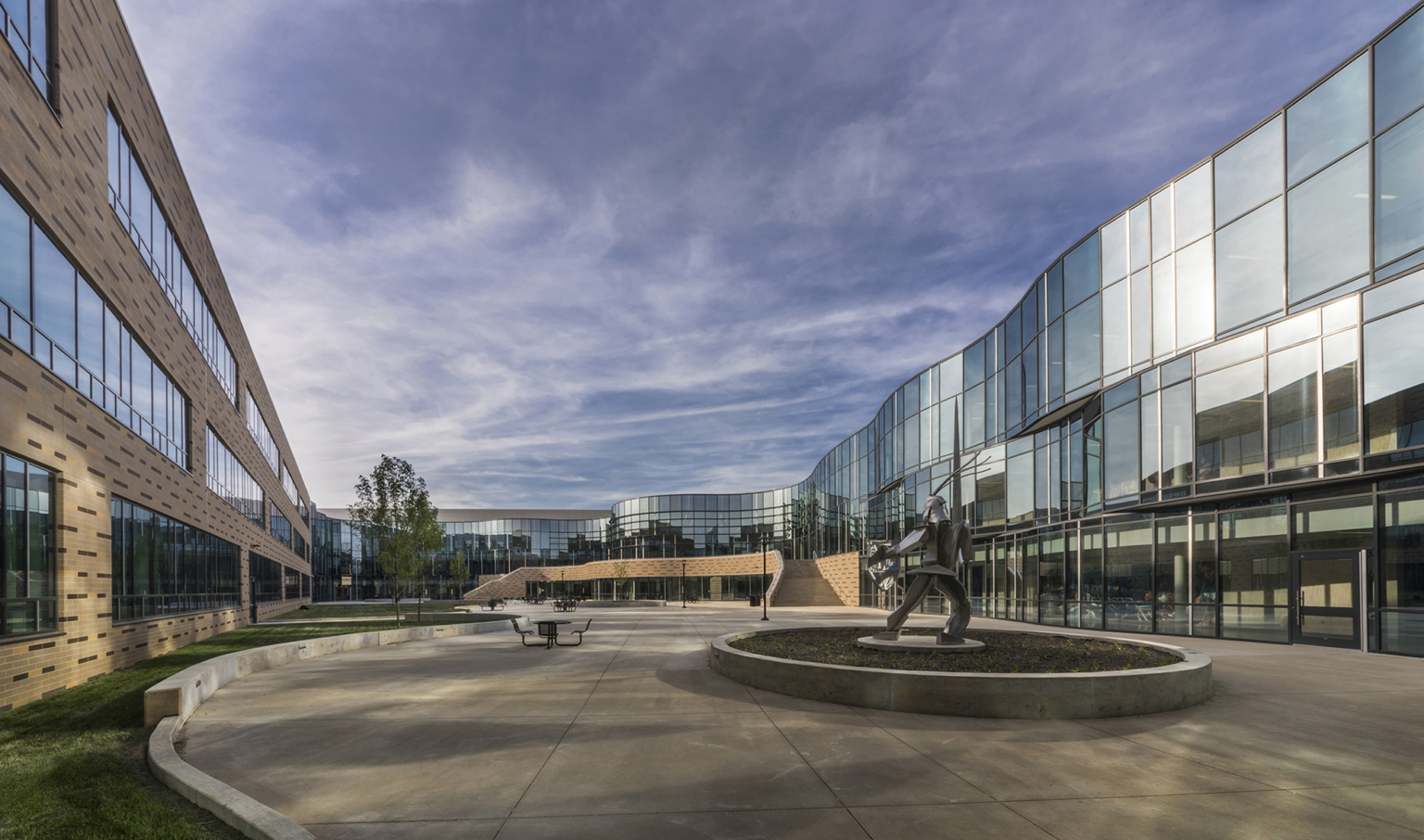 a brick building and mirrored glass facade surround a concrete courtyard with a metal sculpture