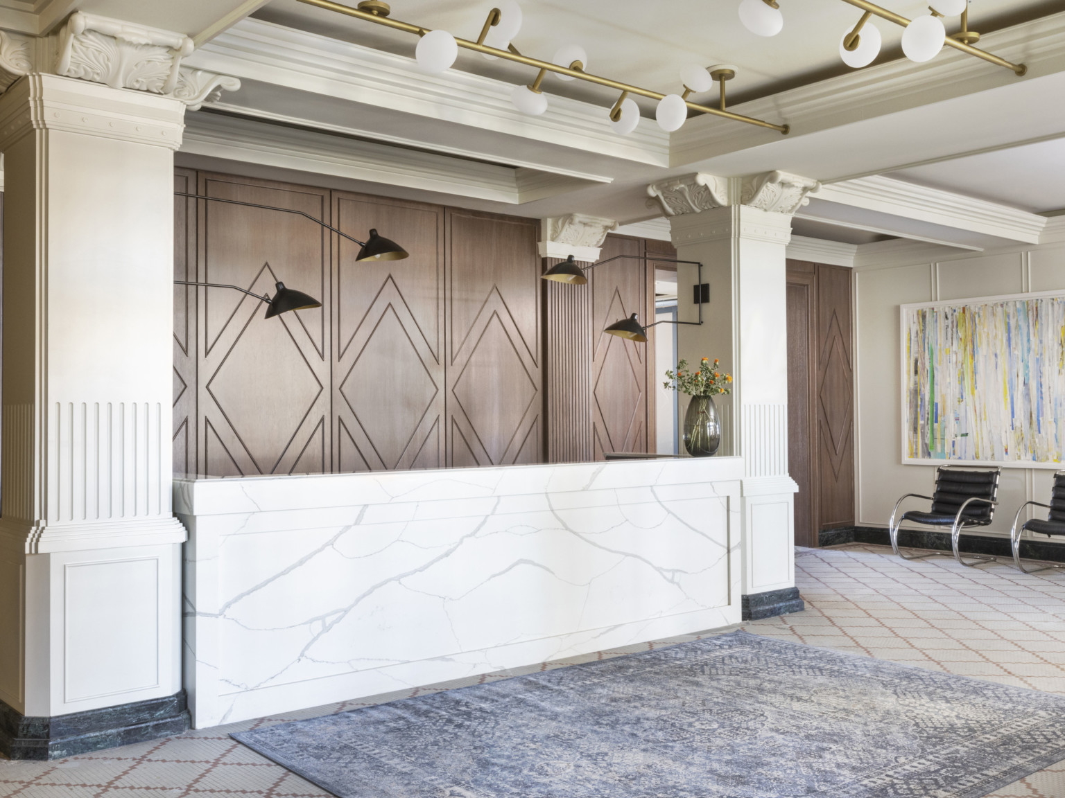 Lobby with white marble reception desk between Corinthian columns in front of geometric wood wall. Blue rug in front of desk