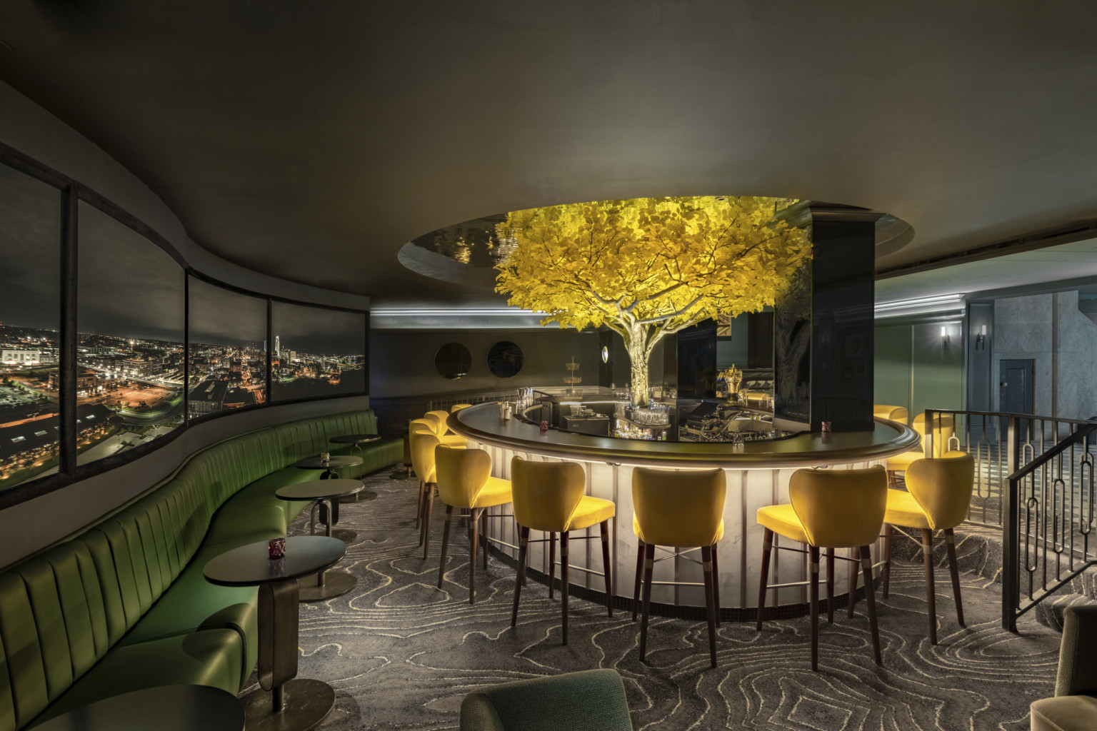 Circular bar with large windows along wall above green velvet booth. Yellow leaved tree in back of bar extending into cut out Circular bar with large windows along wall above green velvet booth. Yellow leaved tree in back of bar extending into cut out
