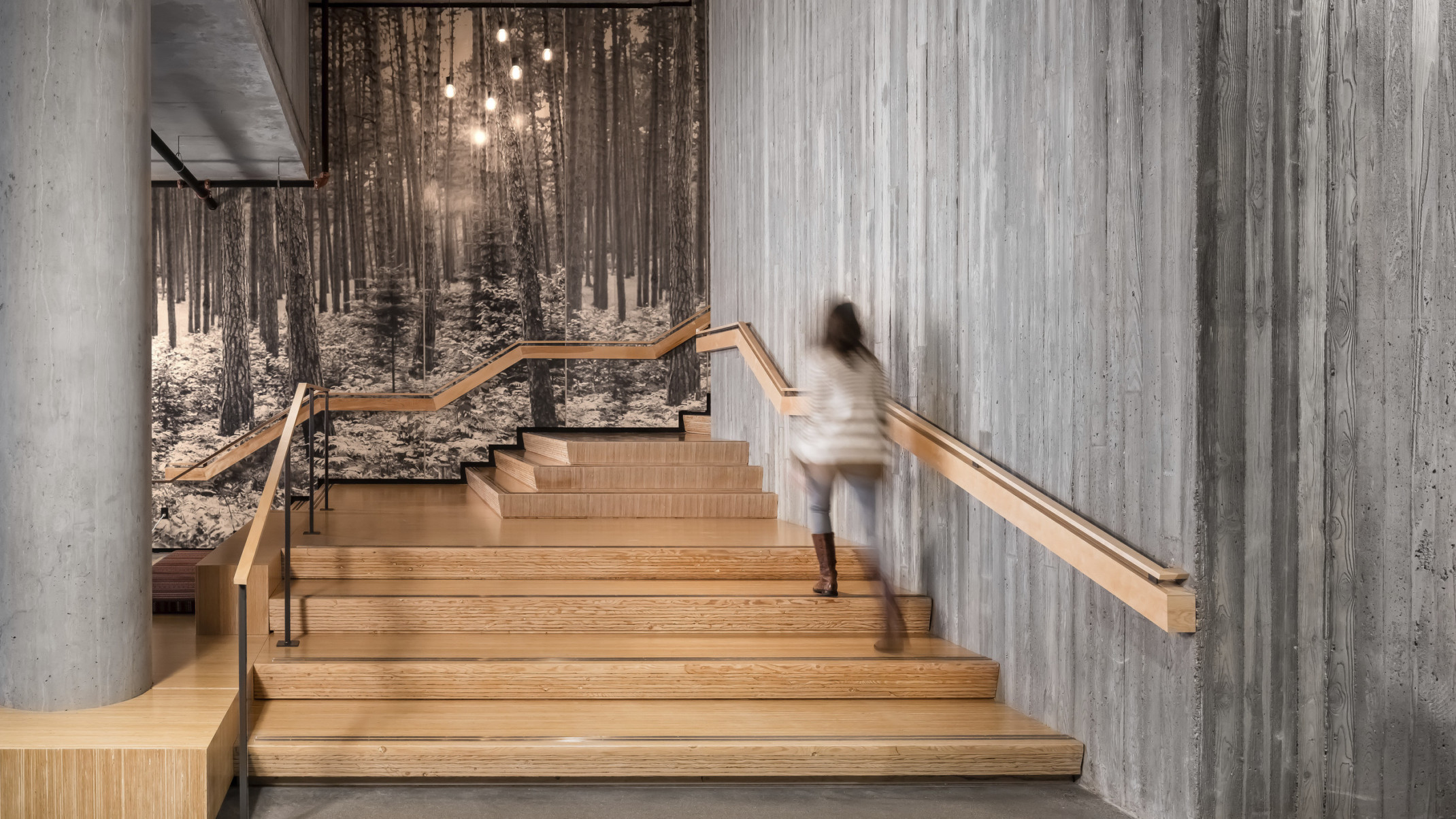 woman walks up wooden stairs surrounded by raw concrete walls. Black and white mural, front of a forest. Angled railing