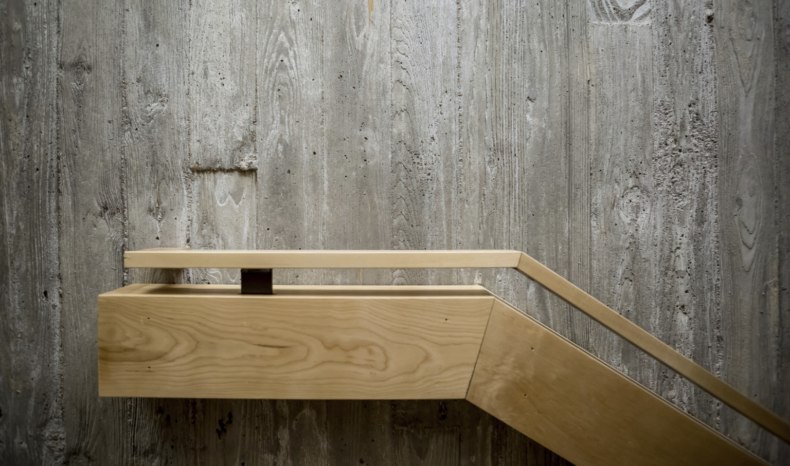 Textural grey raw concrete wall with blonde wood railing angled from right, flattening at center Textural grey raw concrete wall with blonde wood railing angled from right, flattening at center