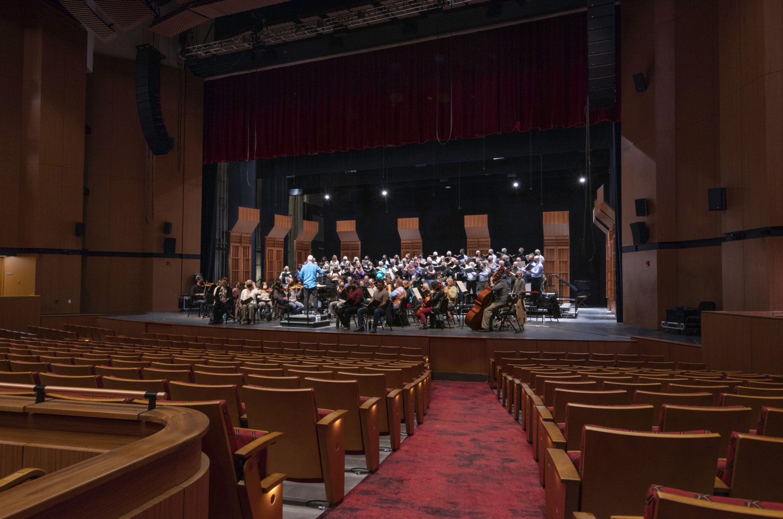 Looking down the isle towards a theater stage where a symphony is playing, surrounded by standalone, operable wood acoustic panels