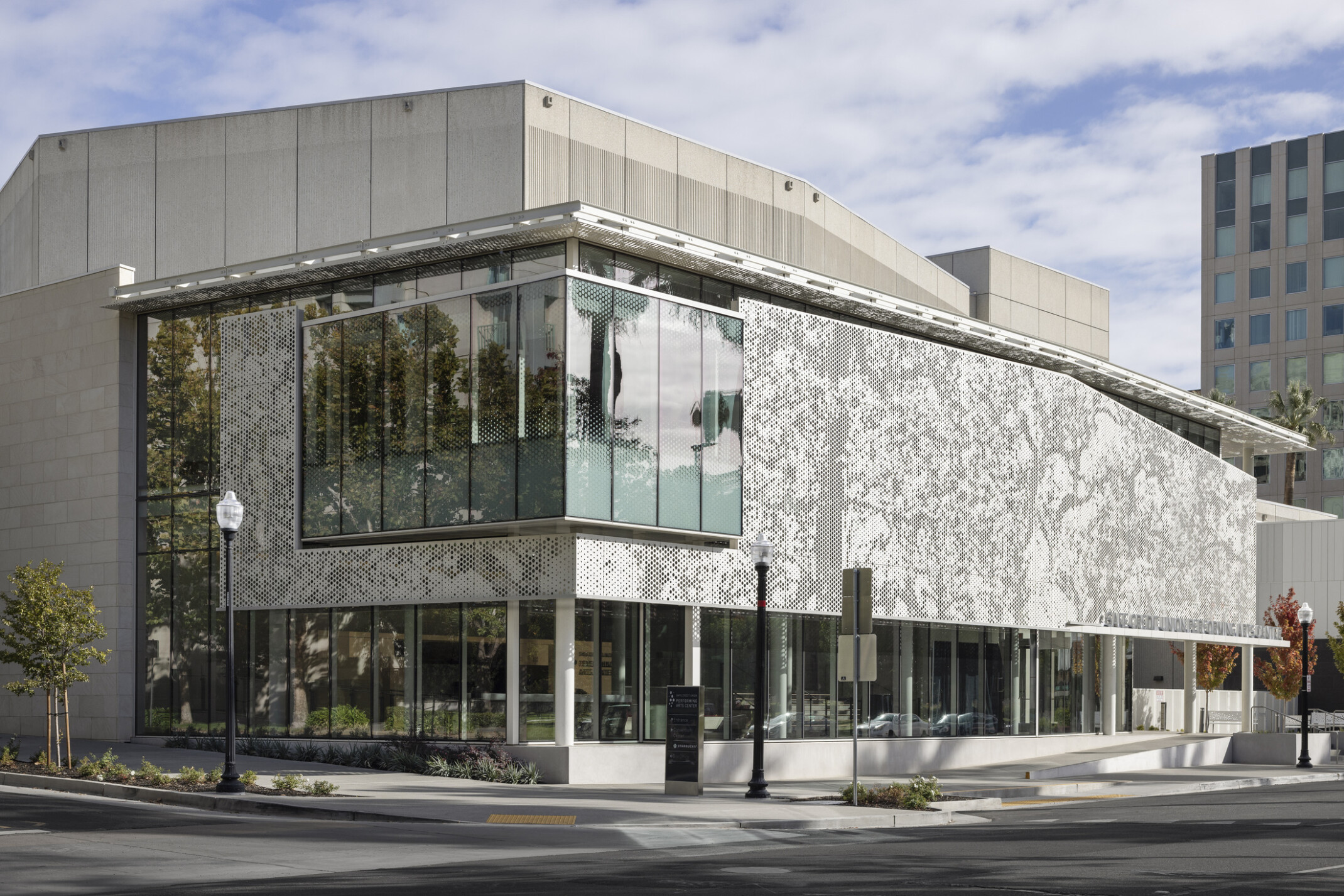 Corner view of a modernized brutalist building, corner glass room hangs over public patio, perforated metal façade provides shade, trees reflect on the glass, scrim