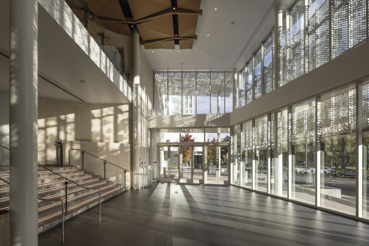Two-story lobby space lit by floor to ceiling glass windows; wood sculpture covers ceiling; perforated light shines through the exterior lacy scrim