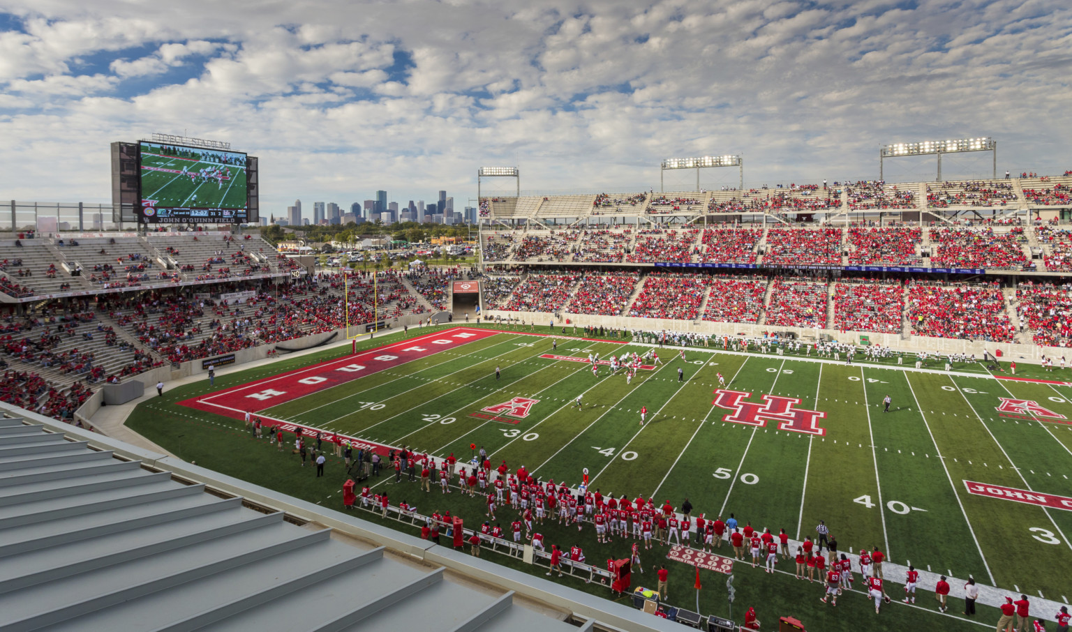 TDECU stadium filled with spectators wearing red and white on game day. Football players on field with UH logo at center