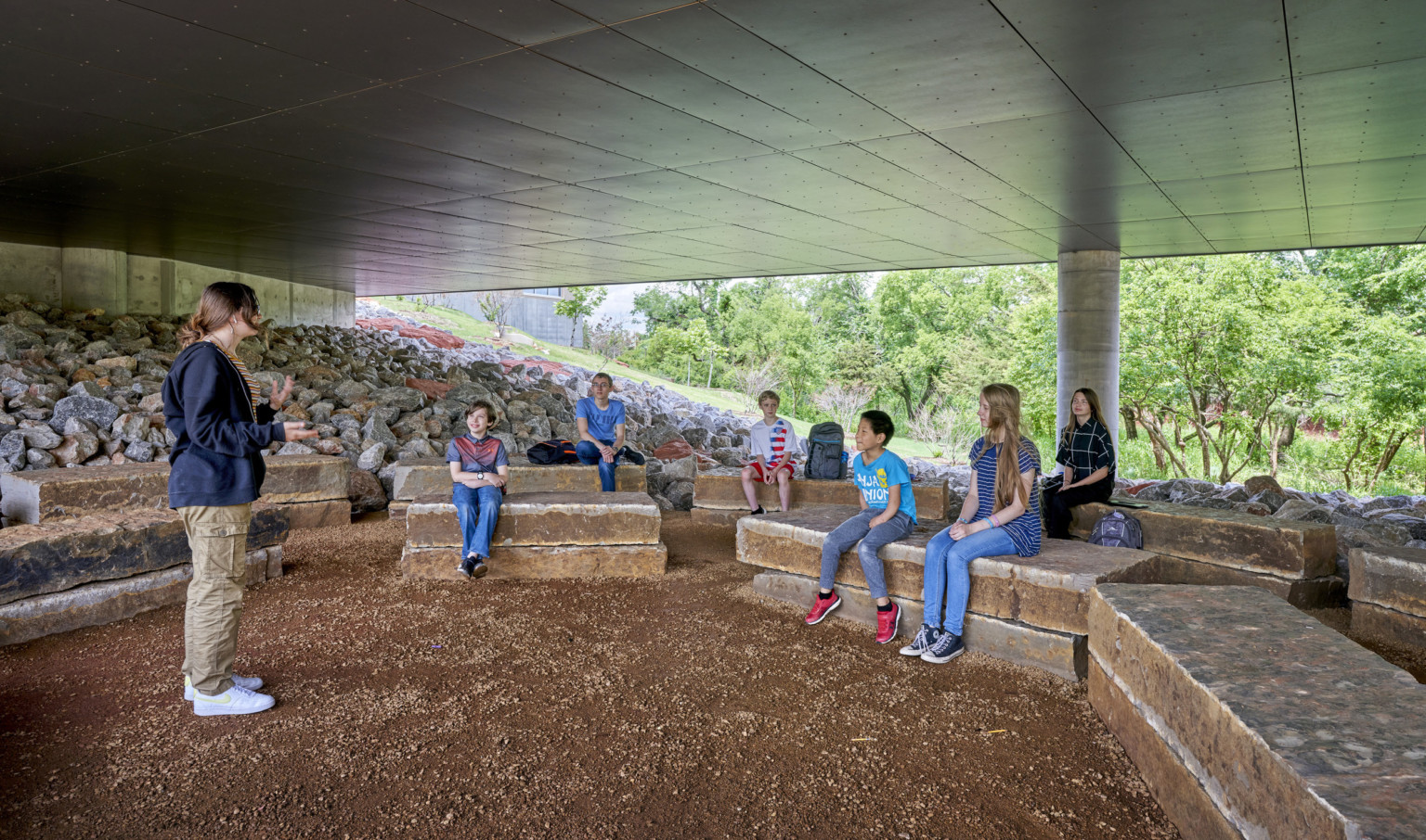 Outdoor learning space with rock benches arranged in semi circle, covered by hallway and open at sides