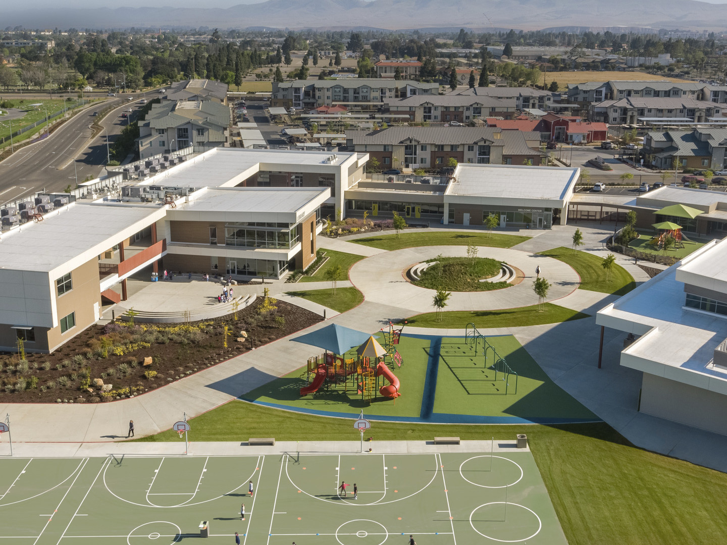 Aerial view from the back of U shaped campus, connected by paths to a circle. Behind, a playground and basketball courts