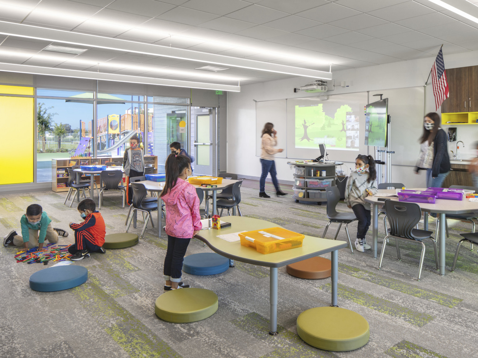 Classroom with tables and floor pillows in front of white board and projector. Left, floor to ceiling window with yellow pane