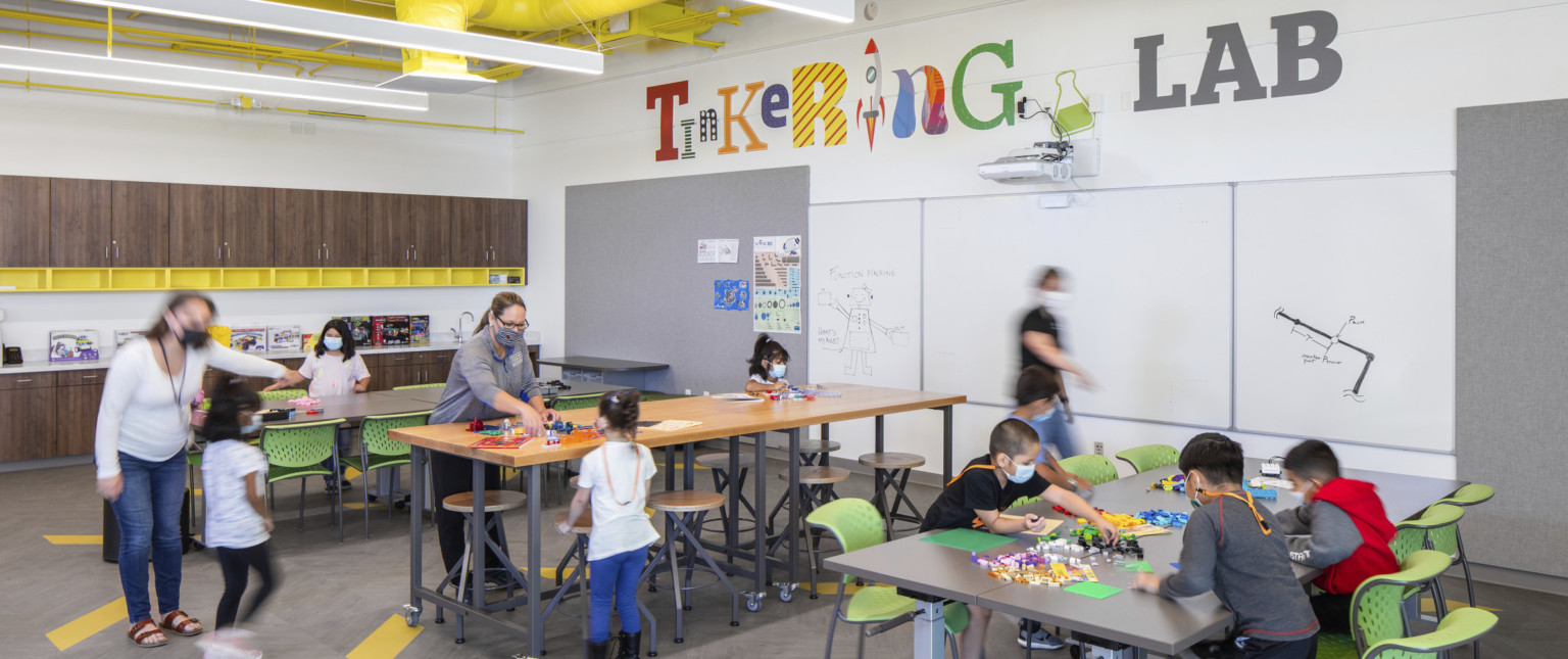 Different heights tables in white room labeled Tinkering Lab. Dark wood cabinets and drawers on left wall, whiteboards front