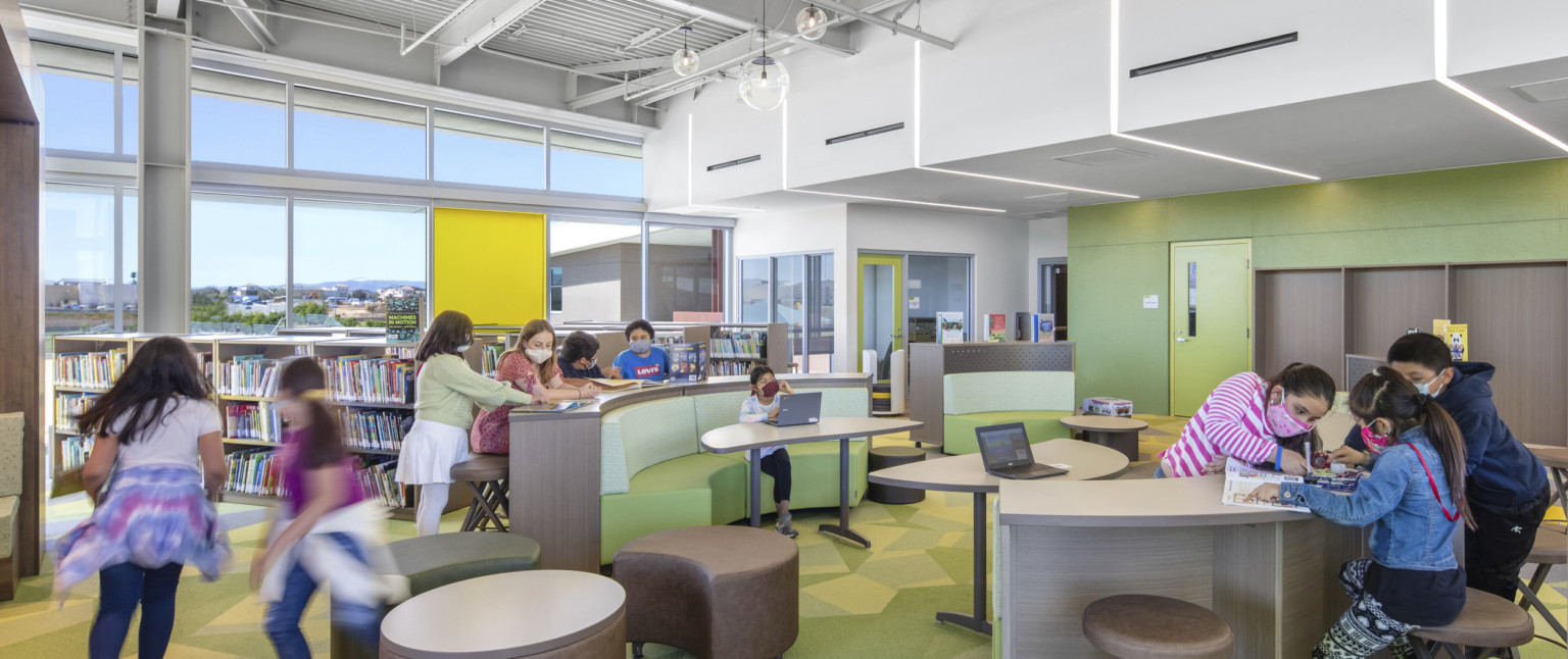 Library with rounded benches and half height book cases. Green accent wall below white drop ceiling. Floor to ceiling window