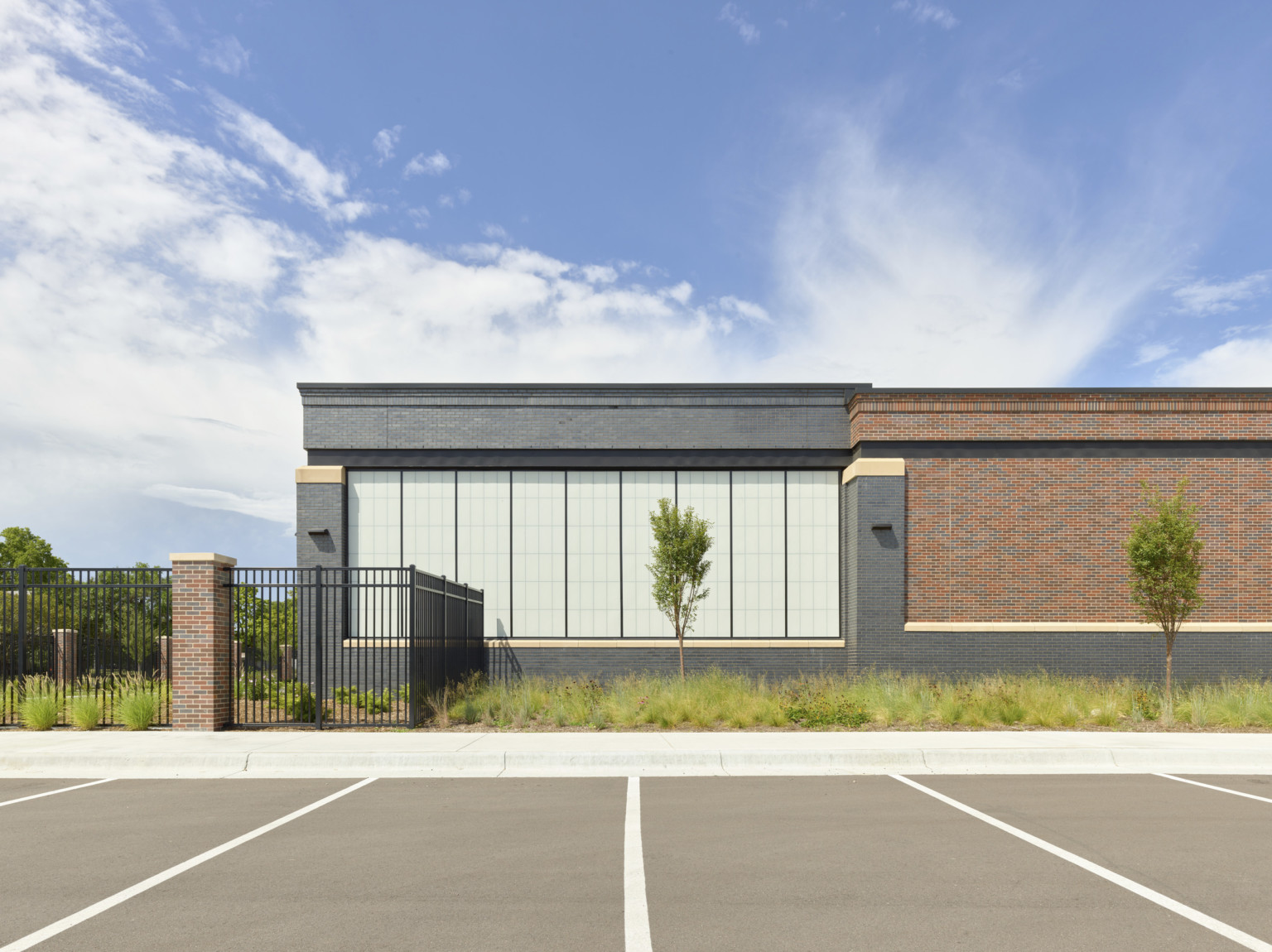 Single story brick building with stone details and large windows. Lawn between building and parking with black metal fence