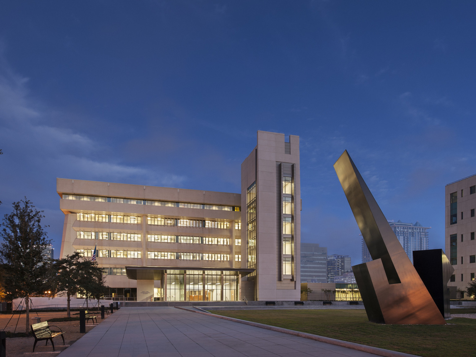 Evening view of multistory George C. Young Federal Building. Light shines out of windows that face walkway and sculpture Evening view of multistory George C. Young Federal Building. Light shines out of windows that face walkway and sculpture