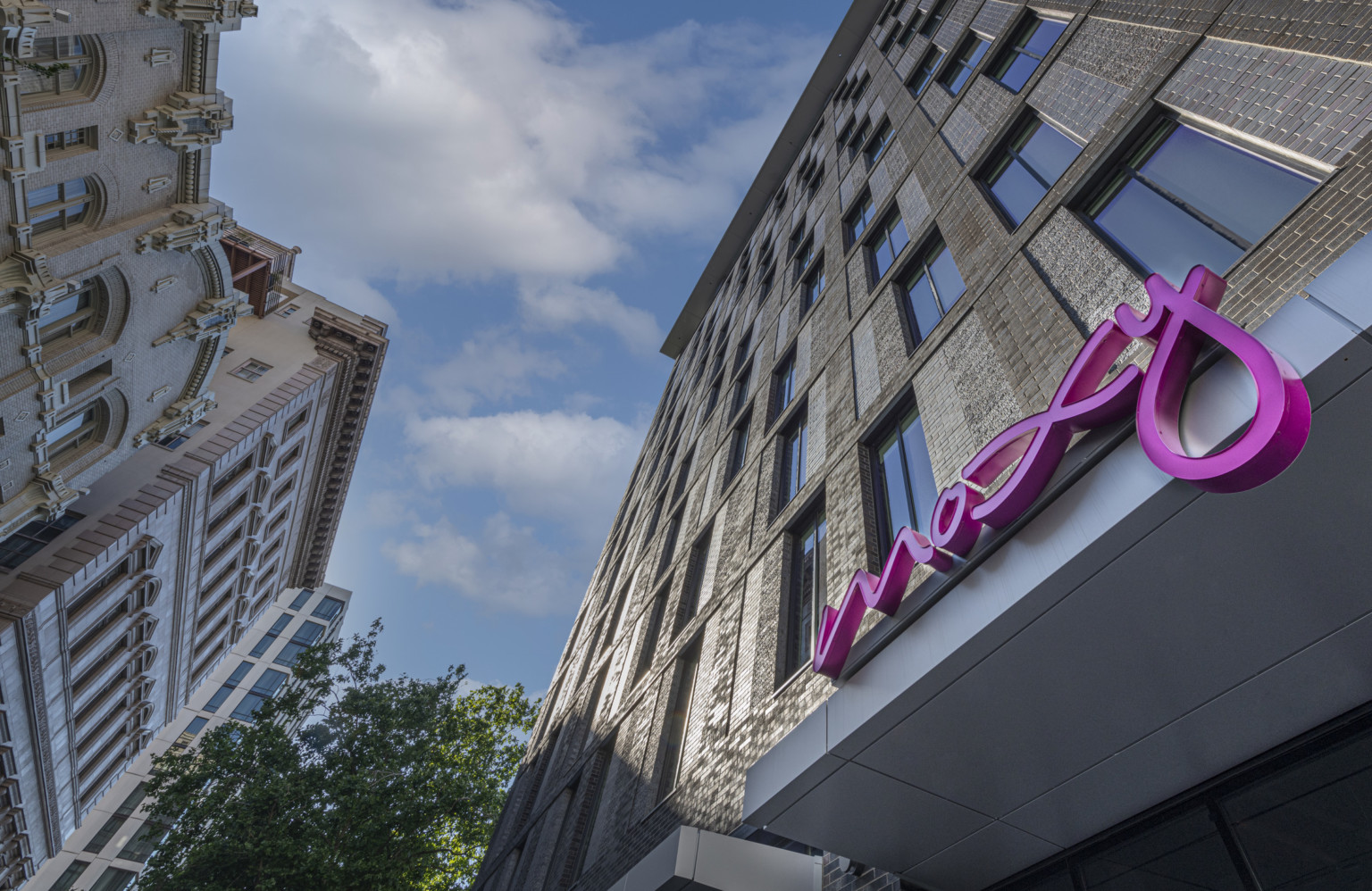 The Moxy Hotel in downtown Portland exterior view looking upwards from the ground at stone building with pink script sign