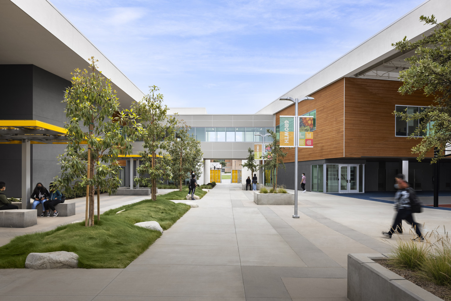 Exterior tree lined walkway between buildings. Grey walls with white at the top, and a wood panel accent on right upper wall