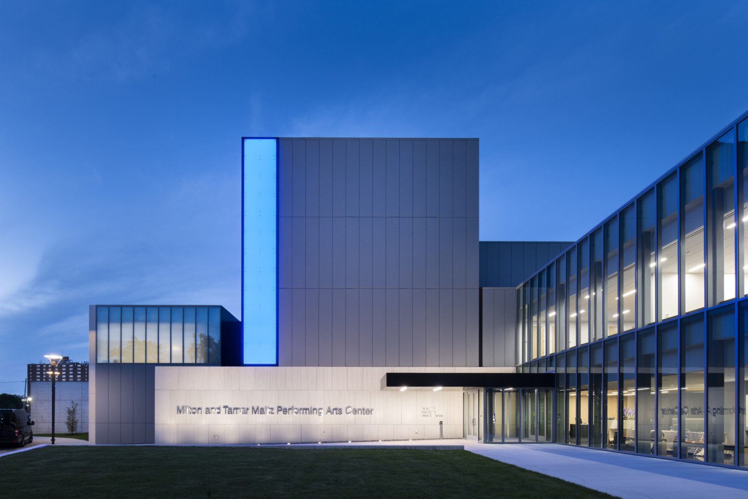 Side view of entry facing concrete wall and sign, grey building extends three stories above with a blue light panel on left