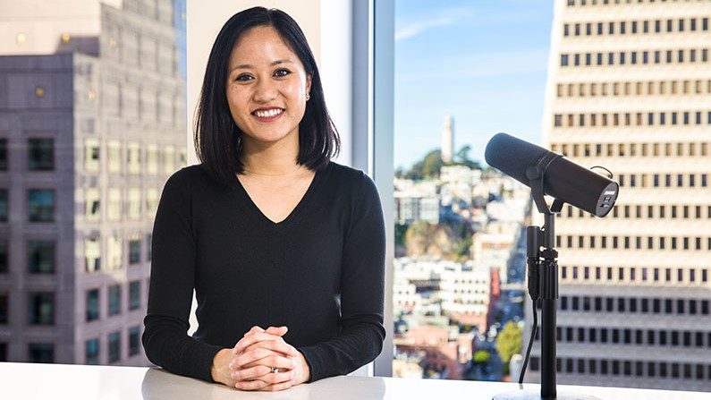 Catherine Meng sits with a microphone pointed at her in front of large windows with a view of high rise buildings