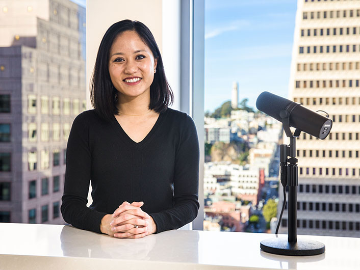 Catherine Meng sits with a microphone pointed at her in front of large windows with a view of high rise buildings