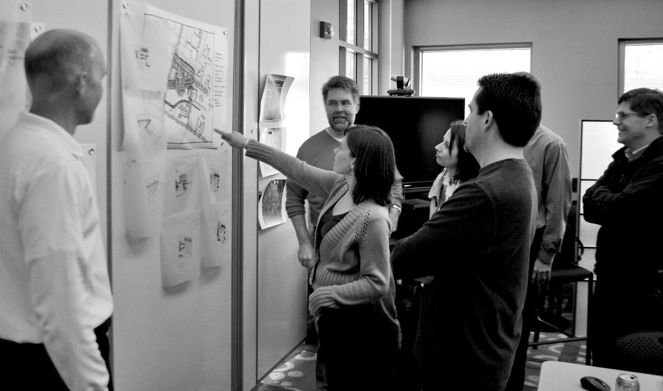 Black and white image of 7 people gathered around site map and other papers hung on wall. Young woman points to image