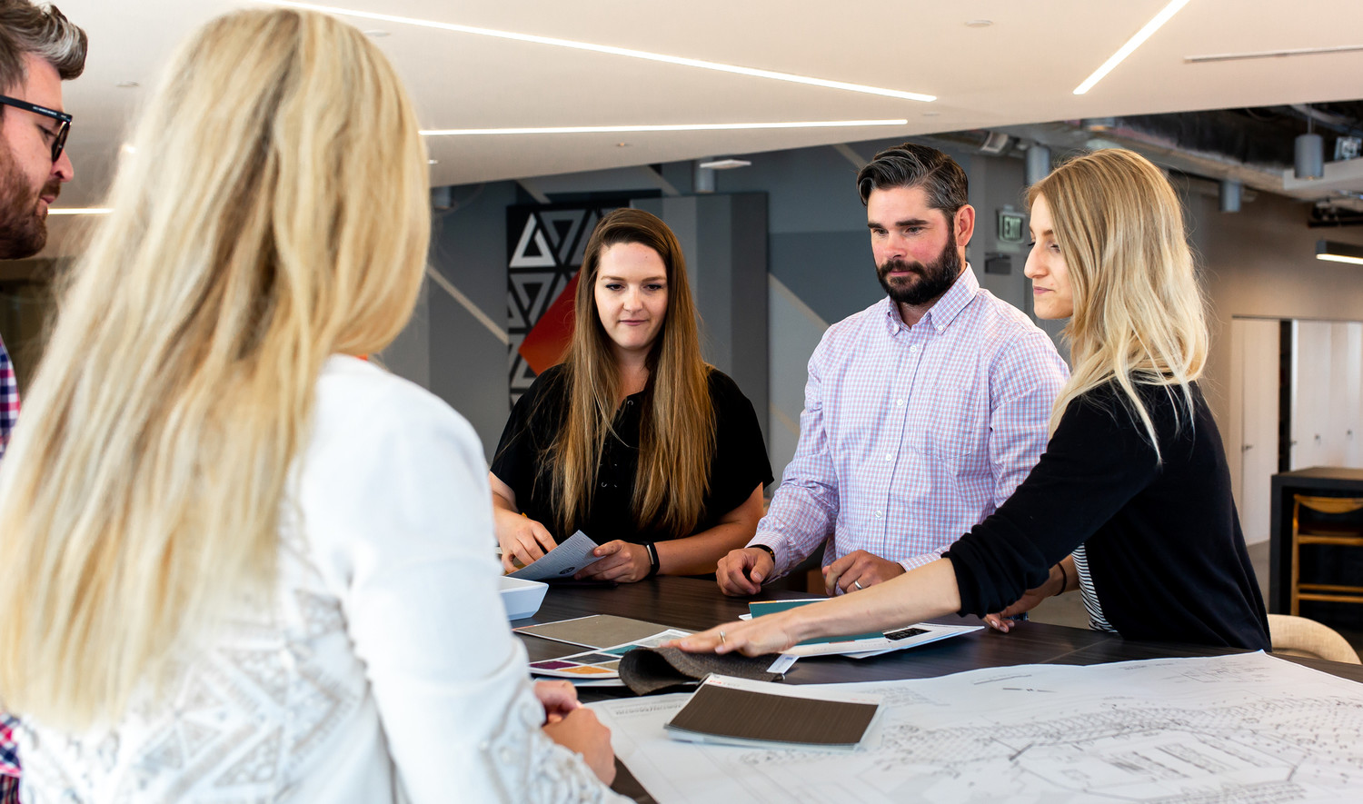 People gathered around standing height dark wood table with fabric samples and plans. Grey wall, back, with geometric details People gathered around standing height dark wood table with fabric samples and plans. Grey wall, back, with geometric details