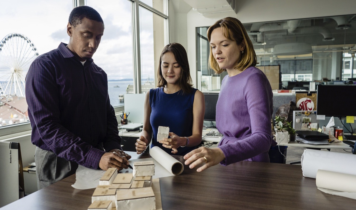 3 young professionals working with stone samples and paper at standing height table by large windows in corner room