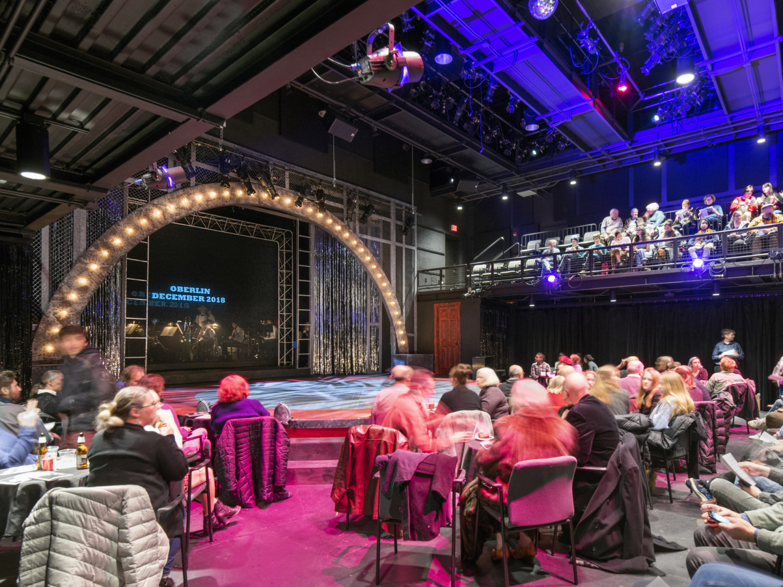 Black box theater audience in flexible first floor seating below balcony facing stage with arch light