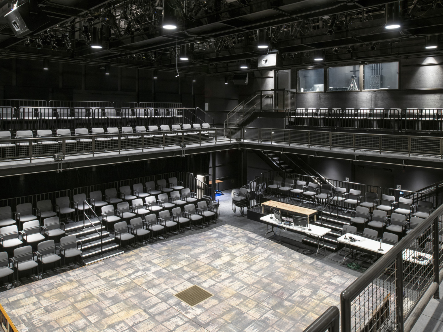 Black box theater with second floor balcony seating, lighting fixtures hang from above. Behind balcony is a room with windows