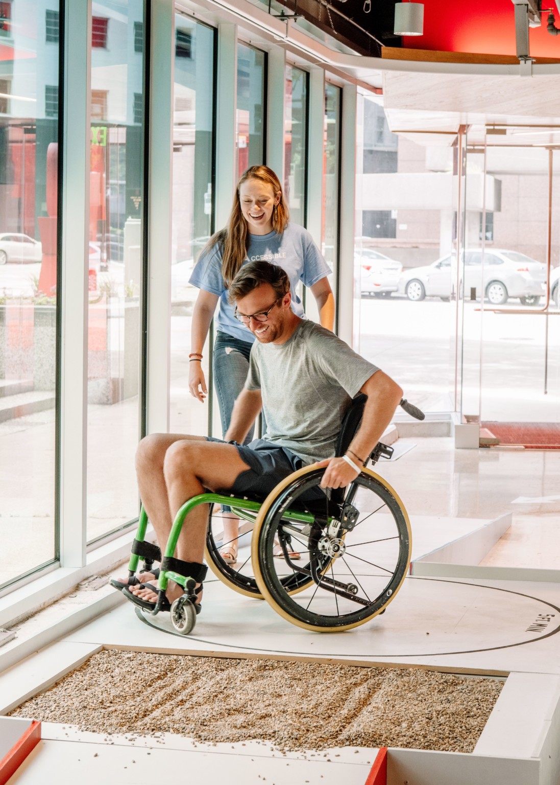 A man sits in wheelchair surrounded by ramp and gravel in room with floor to ceiling windows and red accents