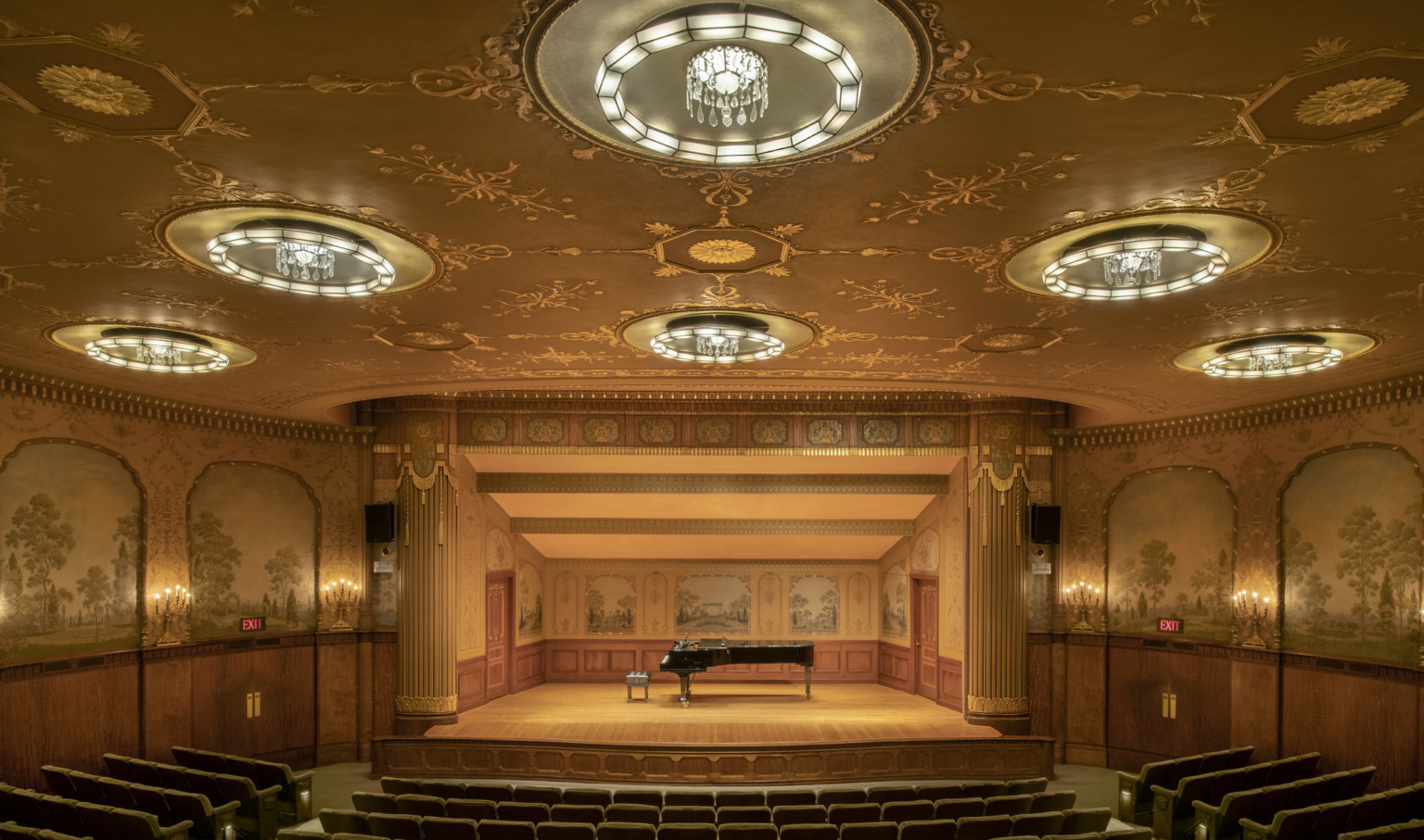 An intimate theater with a gold ceiling and crystal pendant lights. Tapestries and candelabra adorn walls and recital stage.