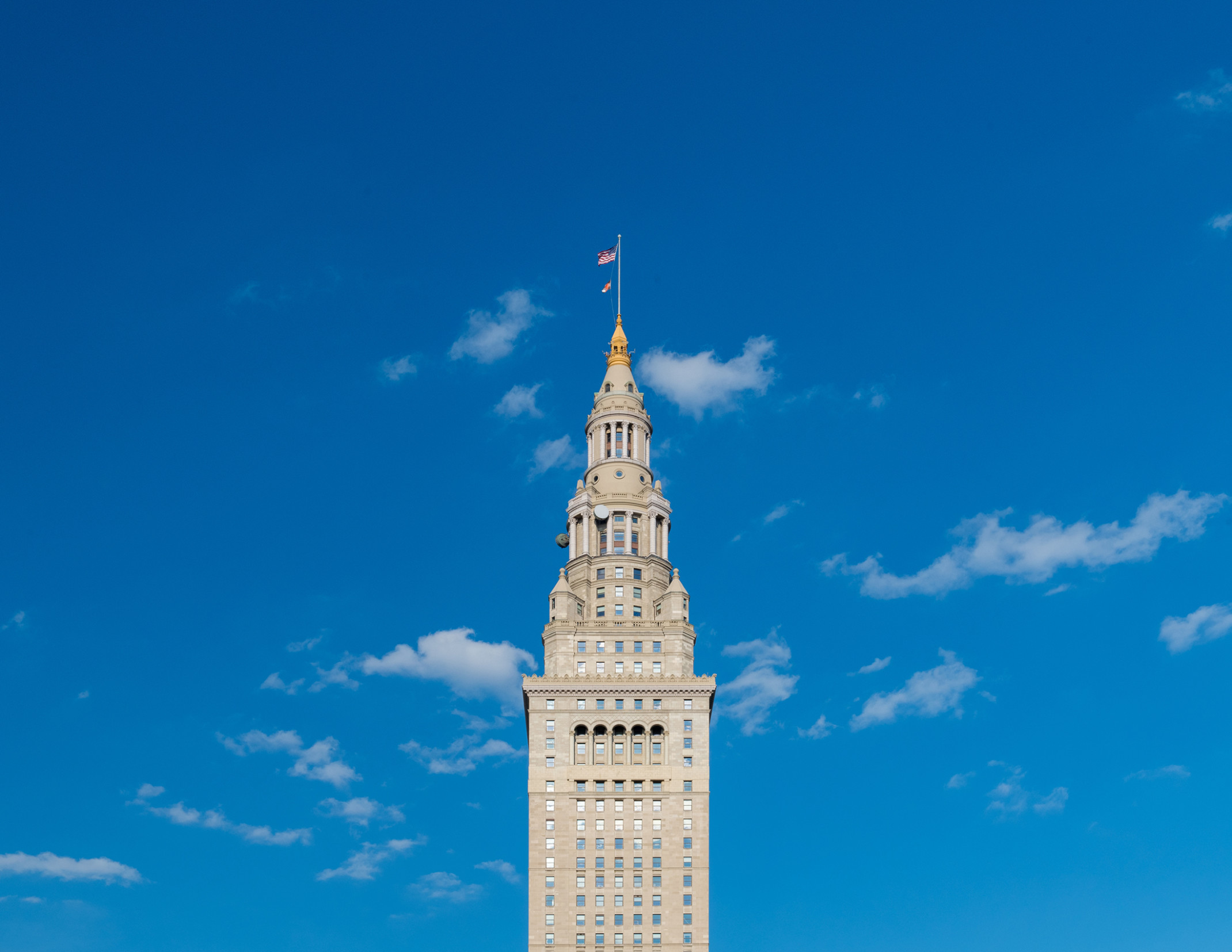 A gray skyscraper with arched windows, columned rotunda, gold cupola, and spire flying a flag on top against a clear blue sky