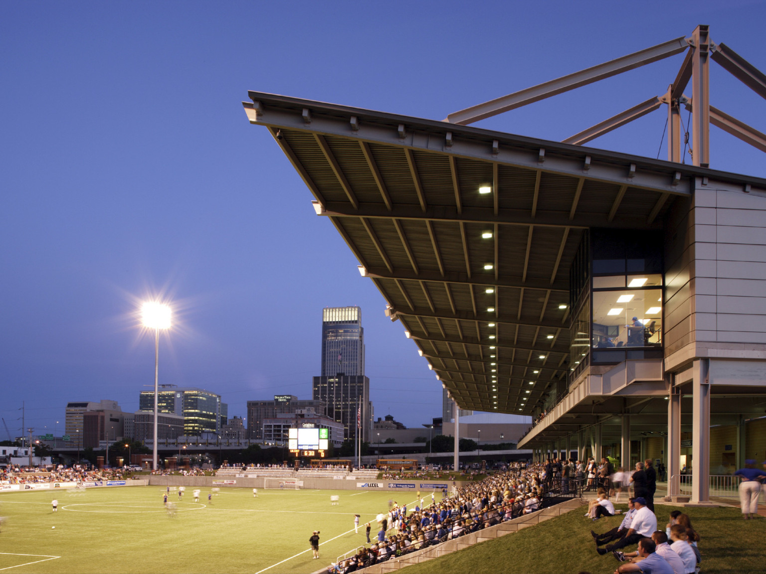 Side view of bleachers with lights in canopy and elevated box seat illuminated from within. Soccer field to left during game