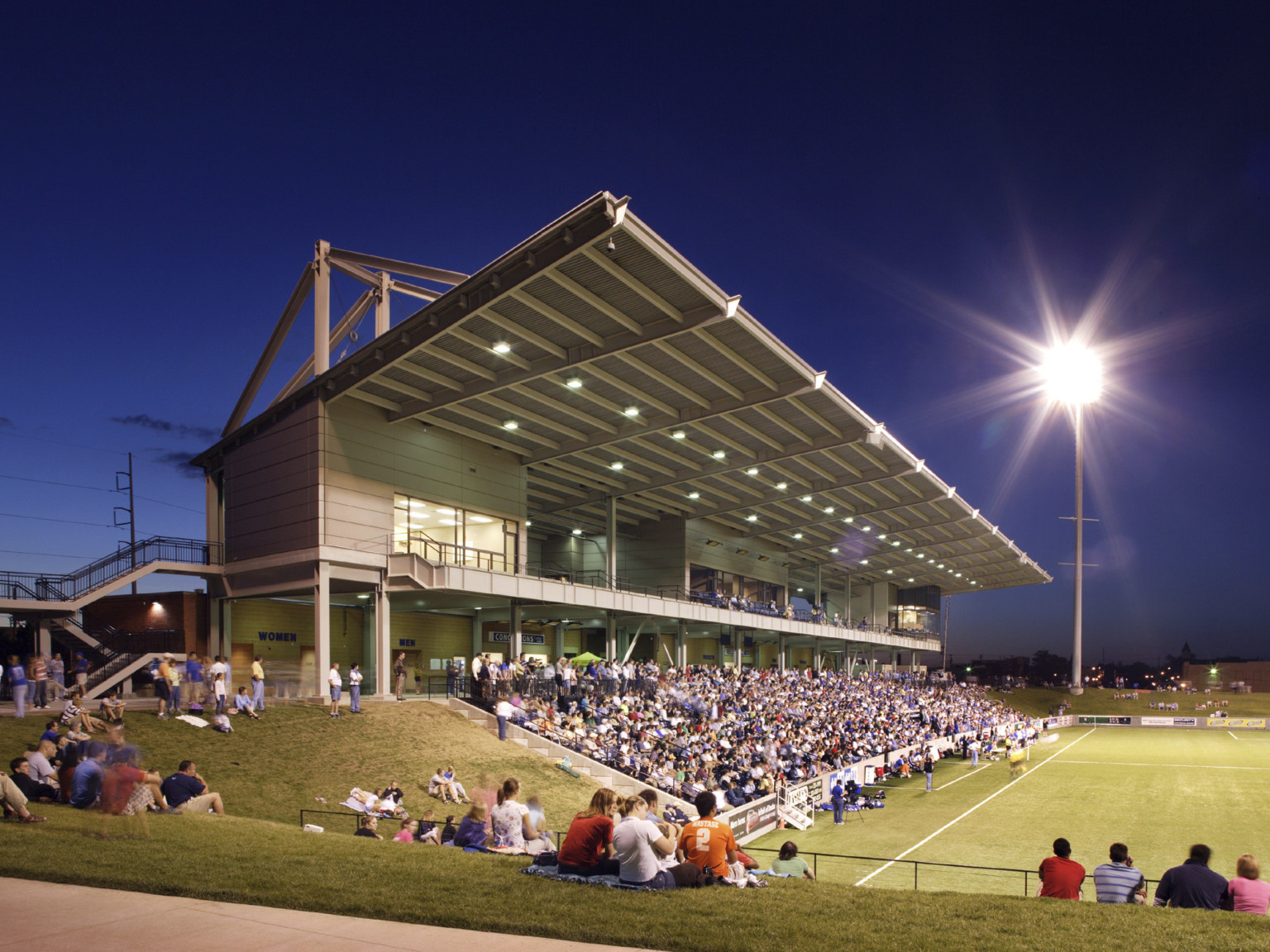 Illuminated canopy extends from roof of elevated grey panel box seating above bleachers. Lawn seating at either end of field