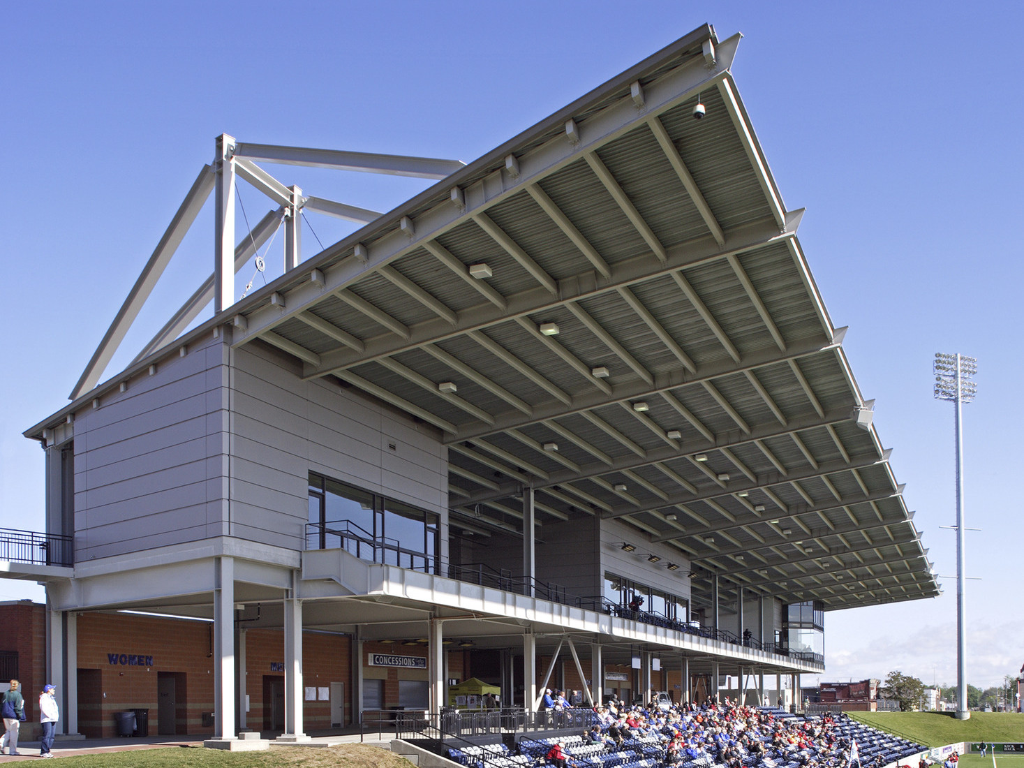 Canopy extends from roof of elevated grey panel box seating above bleachers. Below, brick building with concession stand