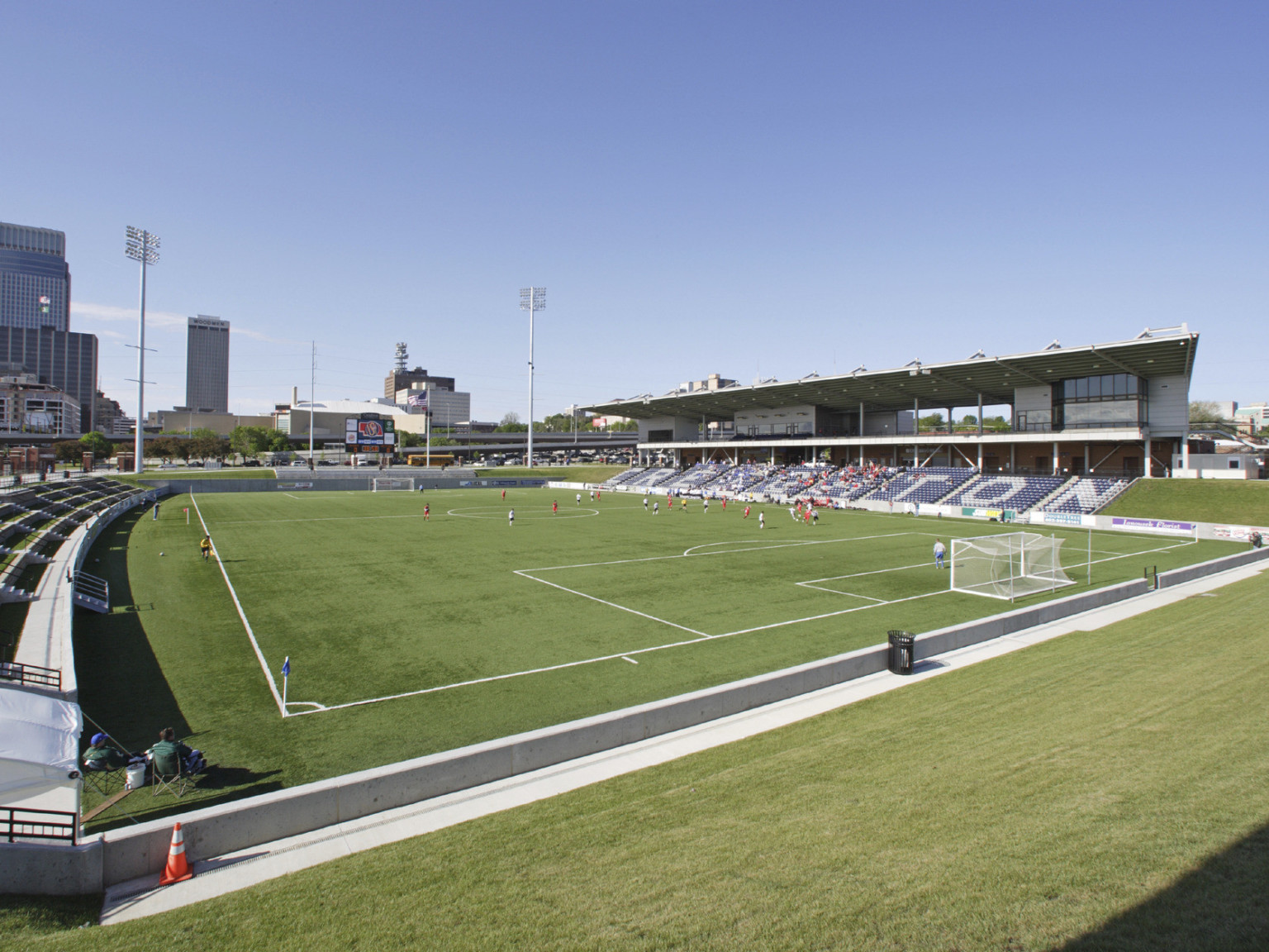 Soccer field viewed from grass lawn end of stadium with sideline seats. Box seats along right sideline, scoreboard at far end